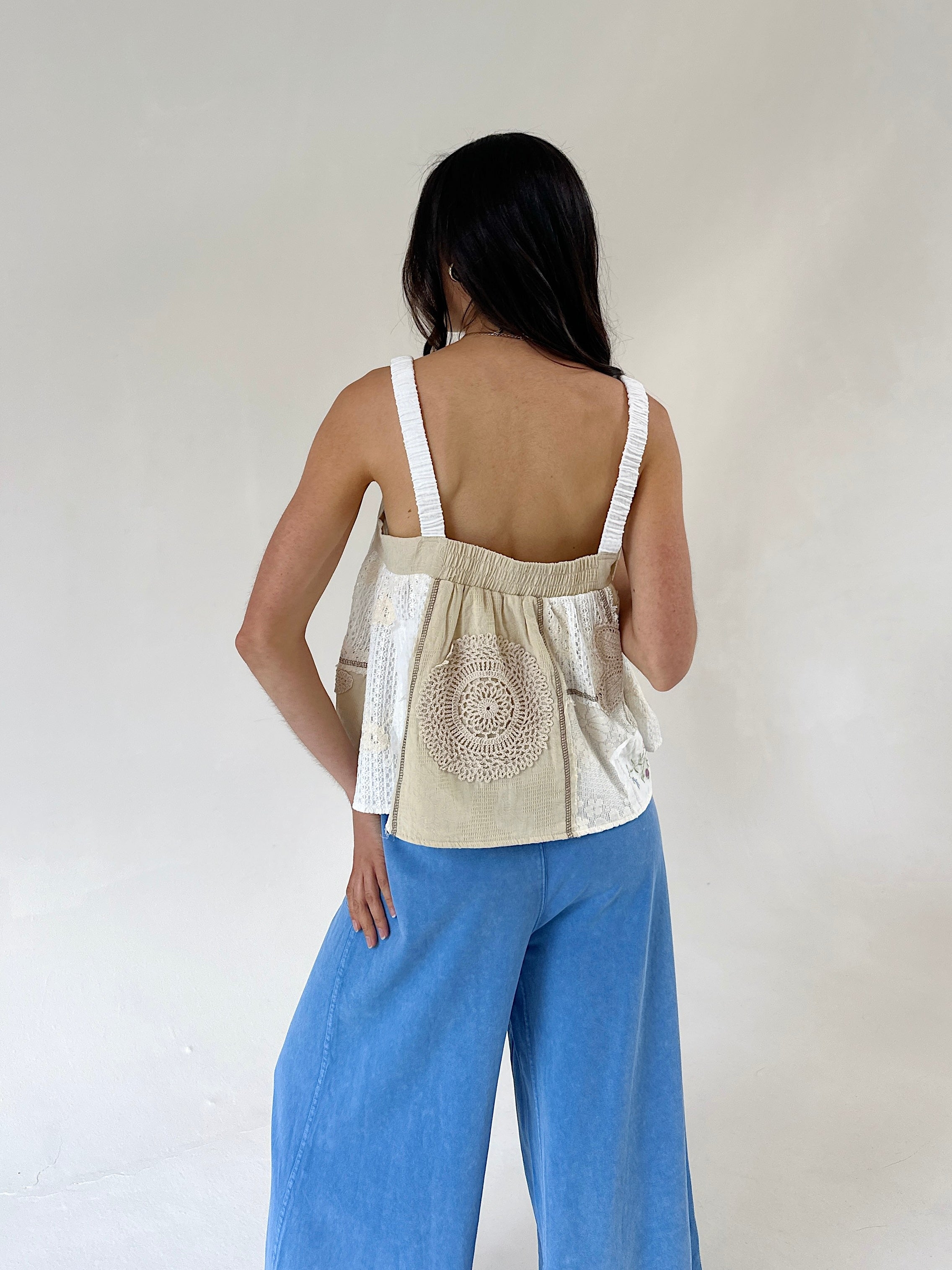 A woman shown from the back wearing a beige sleeveless top with white shoulder straps and a circular embroidered or printed motif on the back, paired with wide-leg blue pants, against a plain off-white studio background.