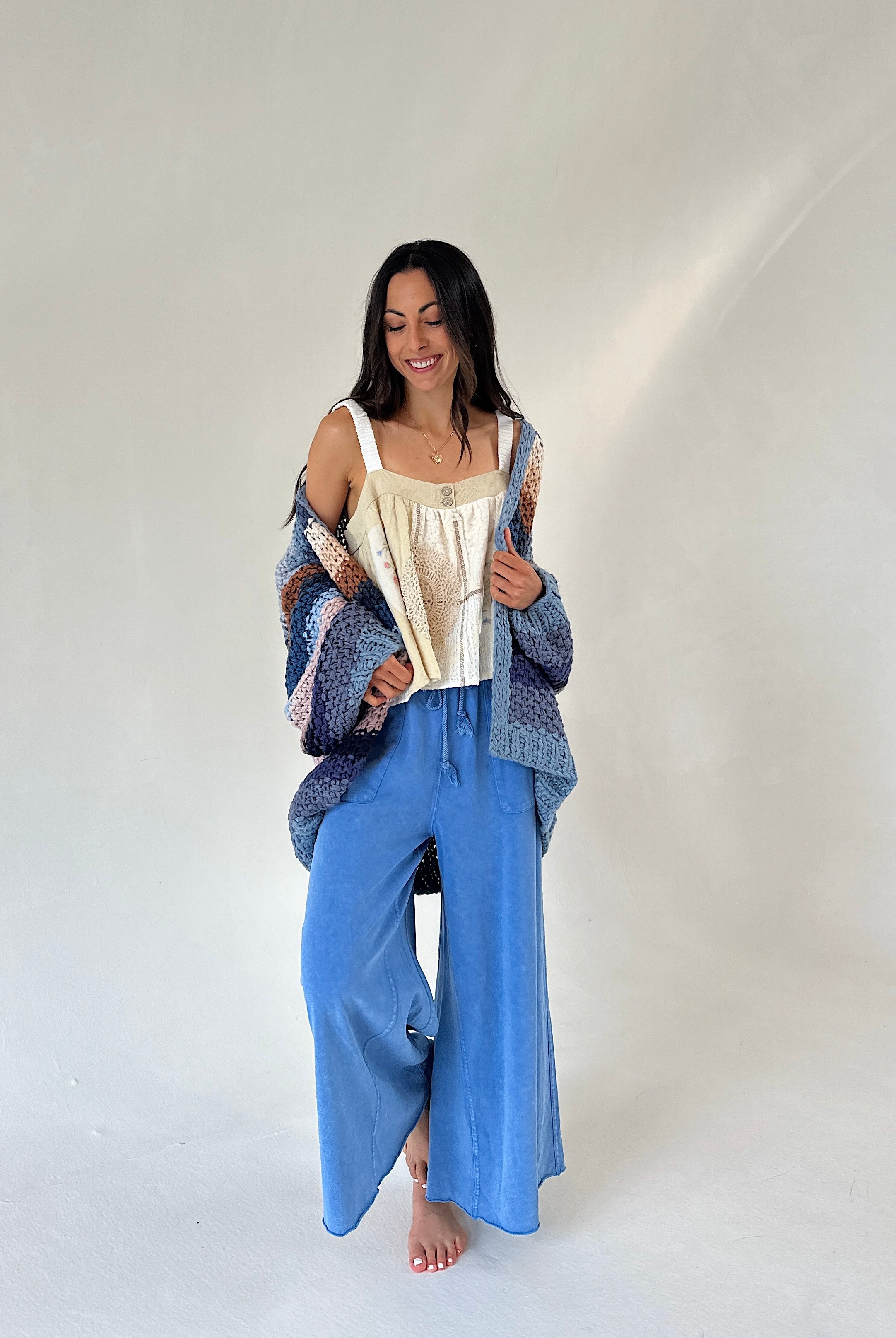 A woman stands barefoot against a plain light studio background, wearing a cream camisole top, wide-leg blue pants, and a loose patterned blue cardigan draped off her shoulders.