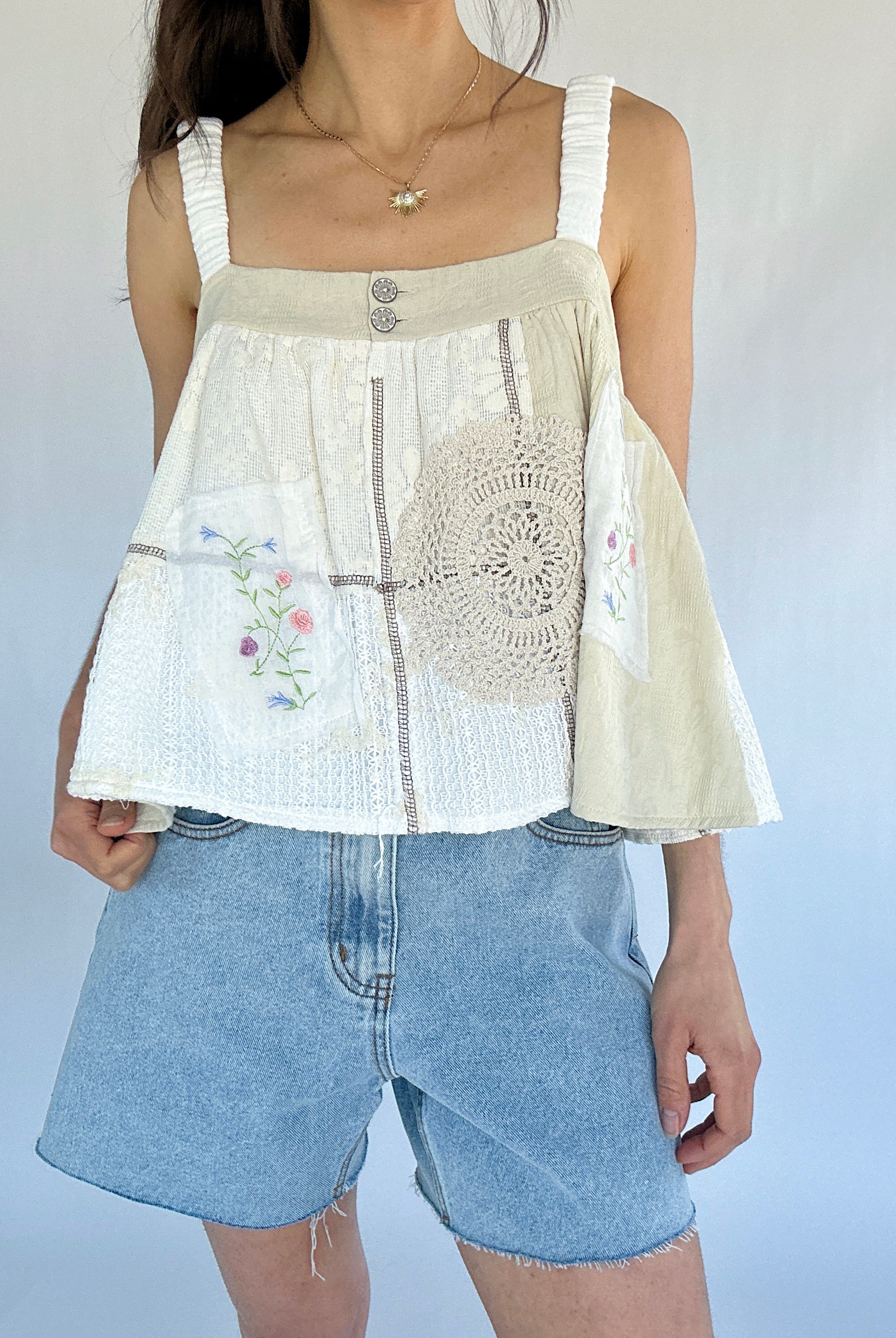 Person wearing a white sleeveless top with crochet and floral embroidered details and denim shorts against a plain background