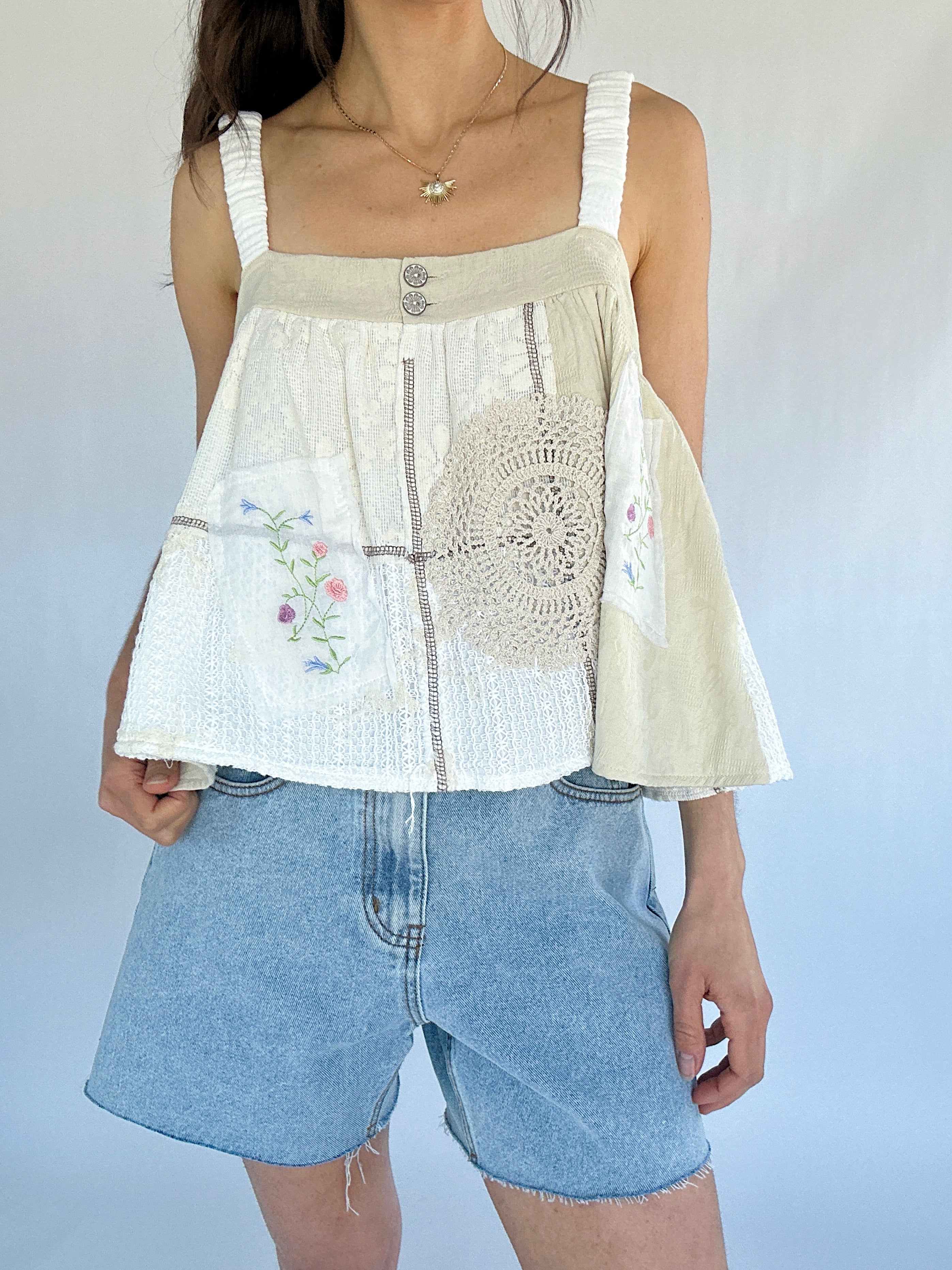 Person wearing a white sleeveless top with crochet and floral embroidered details and denim shorts against a plain background