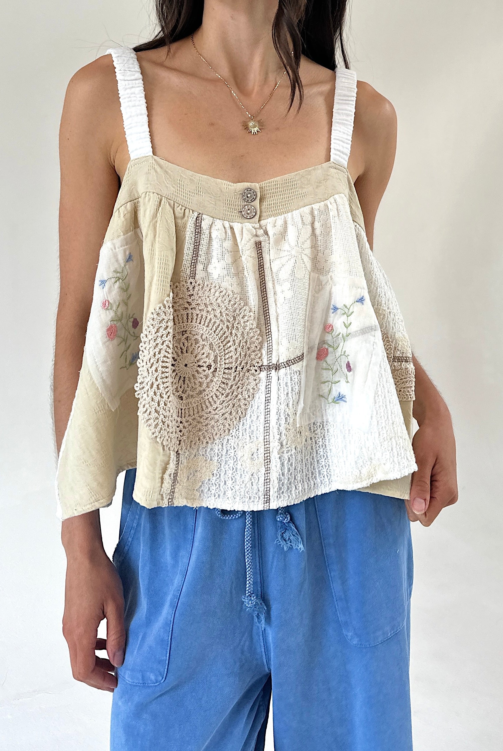 A woman wearing a cream-colored sleeveless top with textured crochet lace panels, floral embroidery, and white shoulder straps, paired with blue drawstring pants against a plain light background.