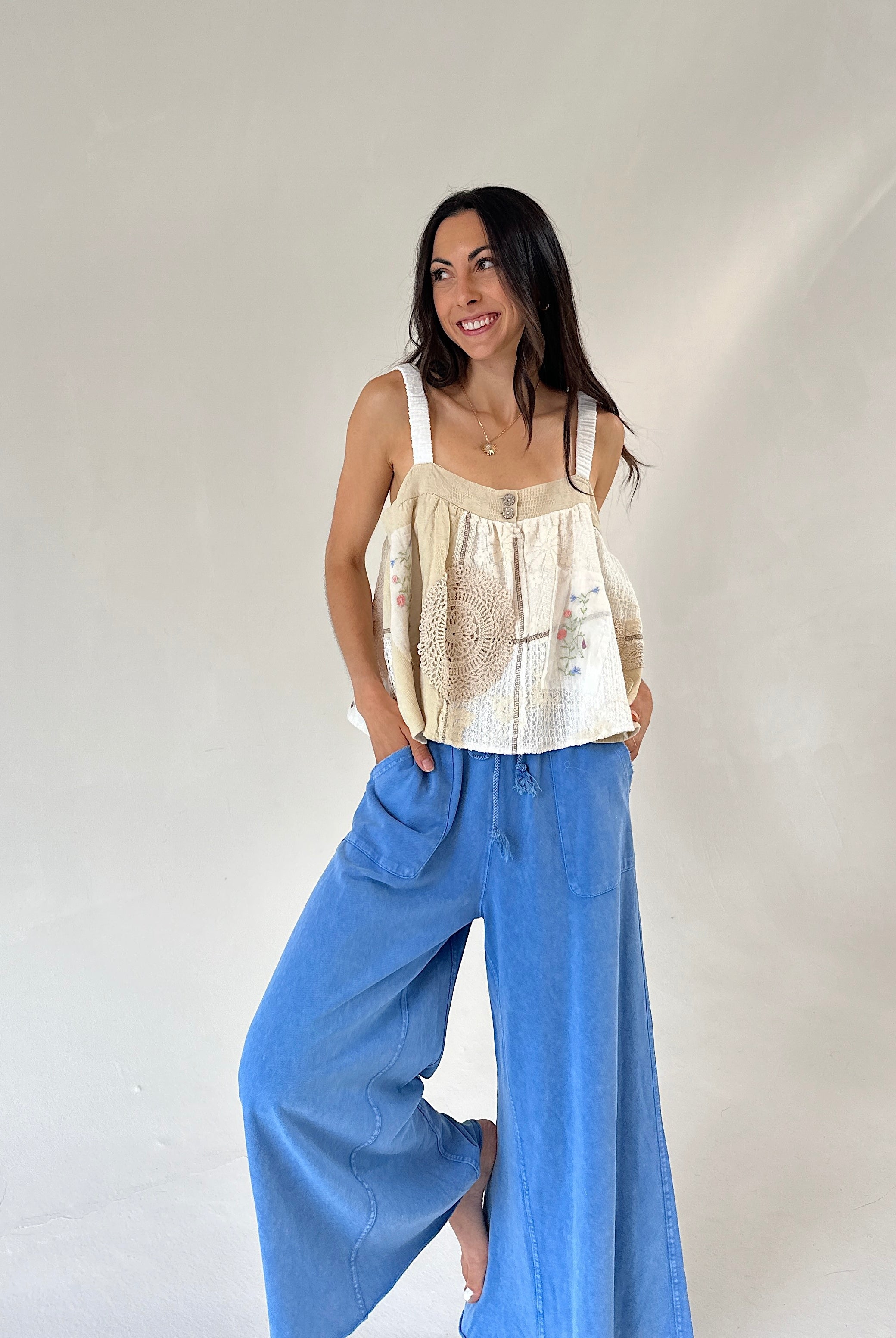A woman modeling a cream sleeveless top with crochet detailing and wide-leg blue jeans against a plain light gray studio background.