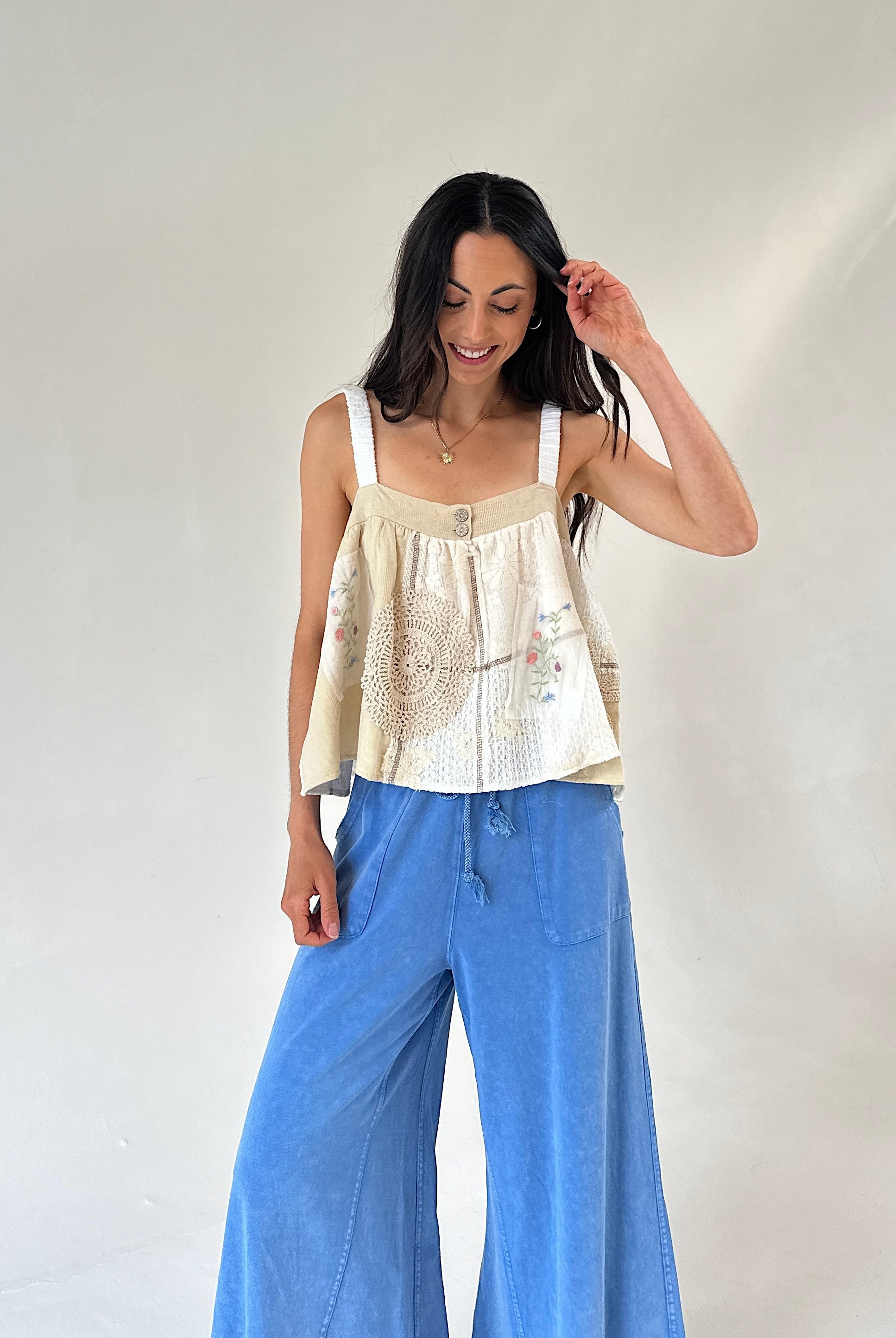 A woman modeling a cream-colored sleeveless top with delicate floral embroidery and wide-leg blue denim pants, standing against a plain off-white studio background.