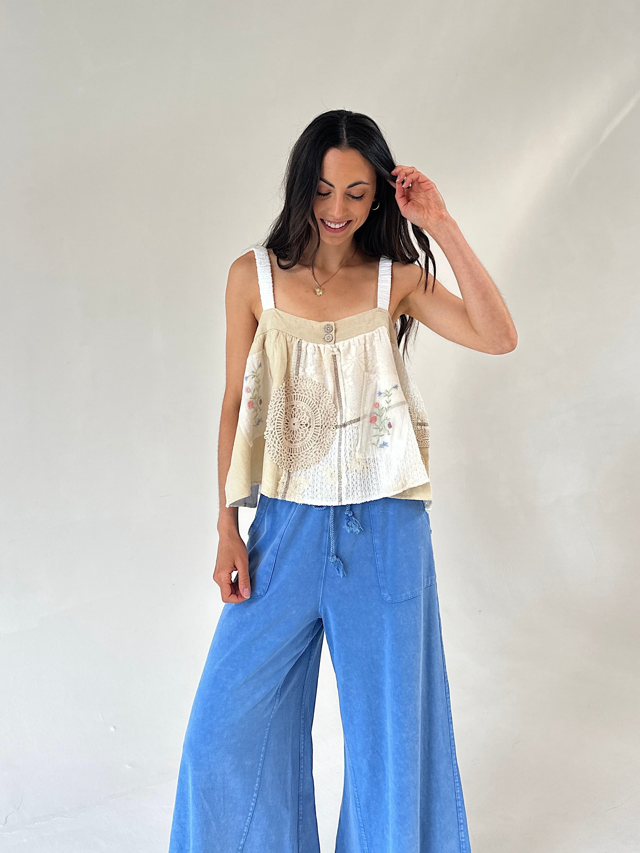 A woman modeling a cream-colored sleeveless top with delicate floral embroidery and wide-leg blue denim pants, standing against a plain off-white studio background.