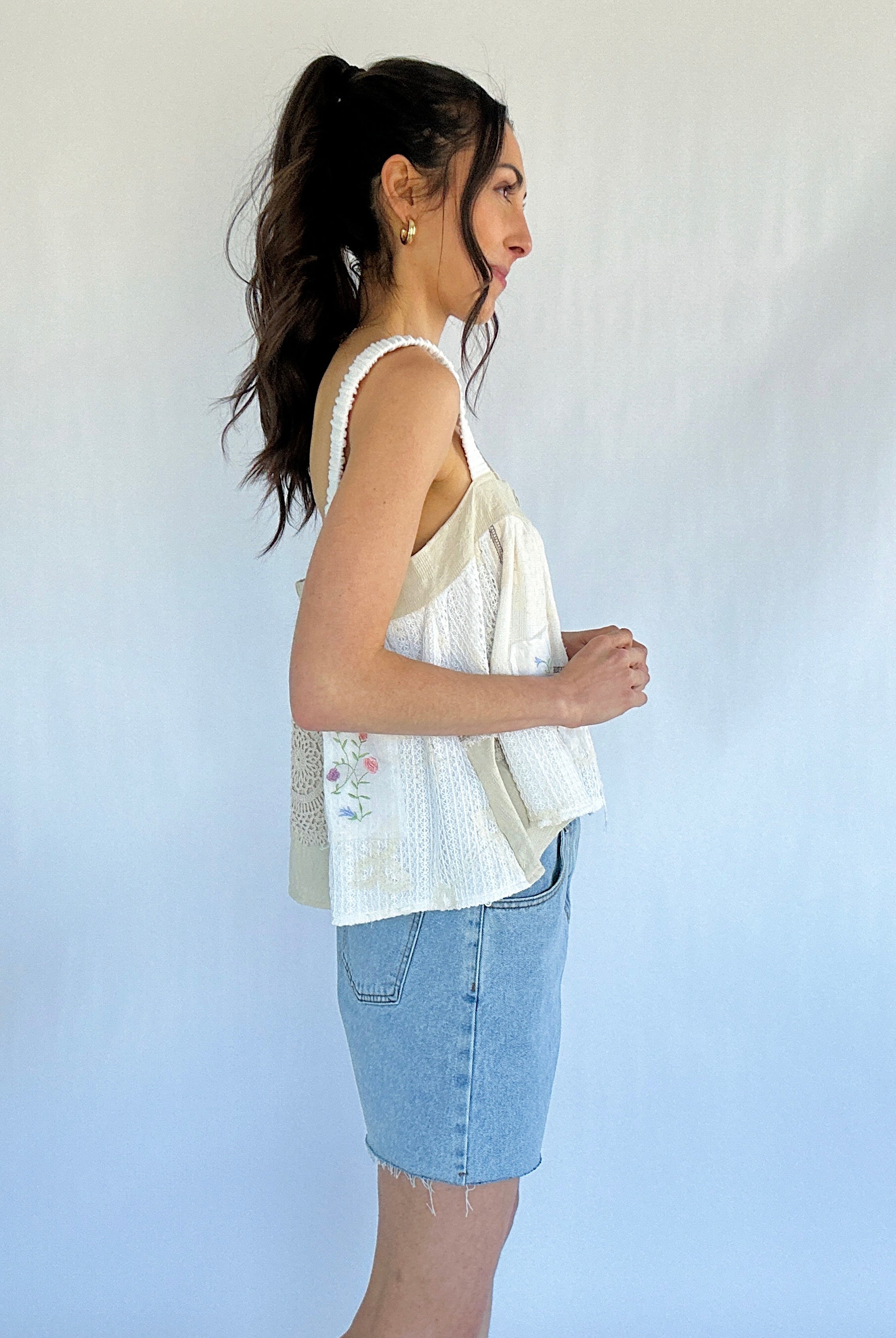 Woman wearing a white sleeveless top and denim shorts against a plain background from side