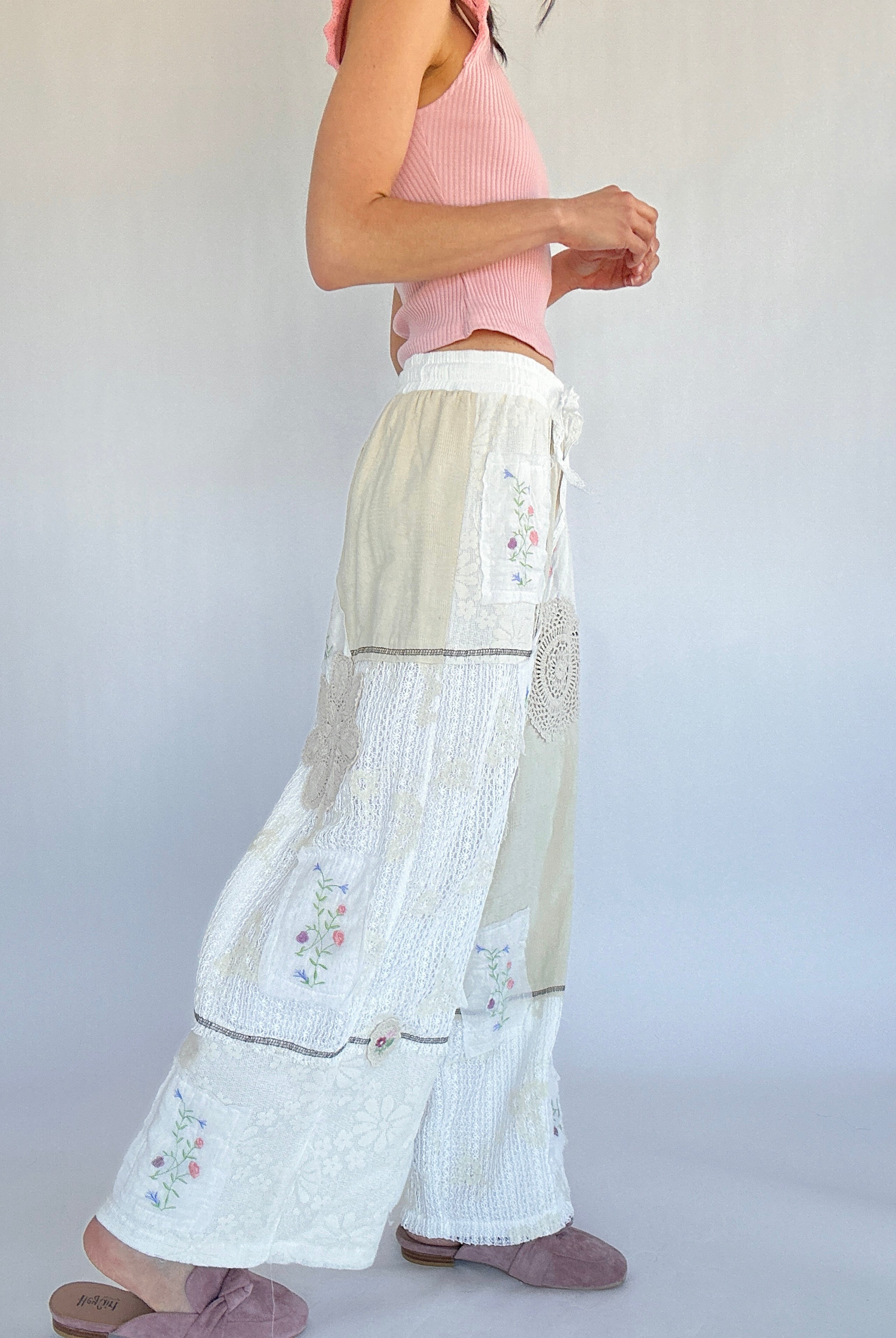 Person wearing a pink top and white pants with floral embroidery on a plain background from side