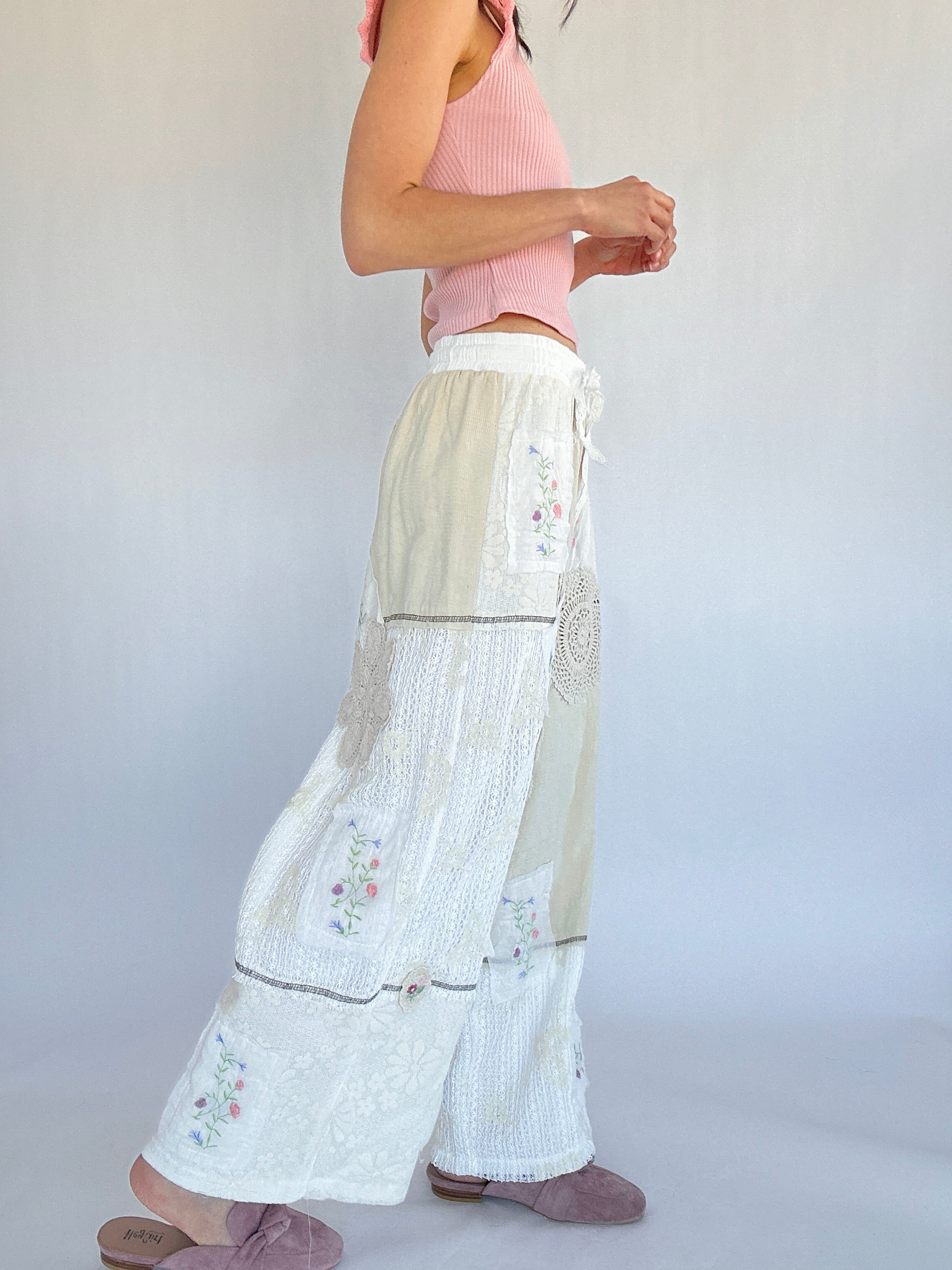 Person wearing a pink top and white pants with floral embroidery on a plain background from side