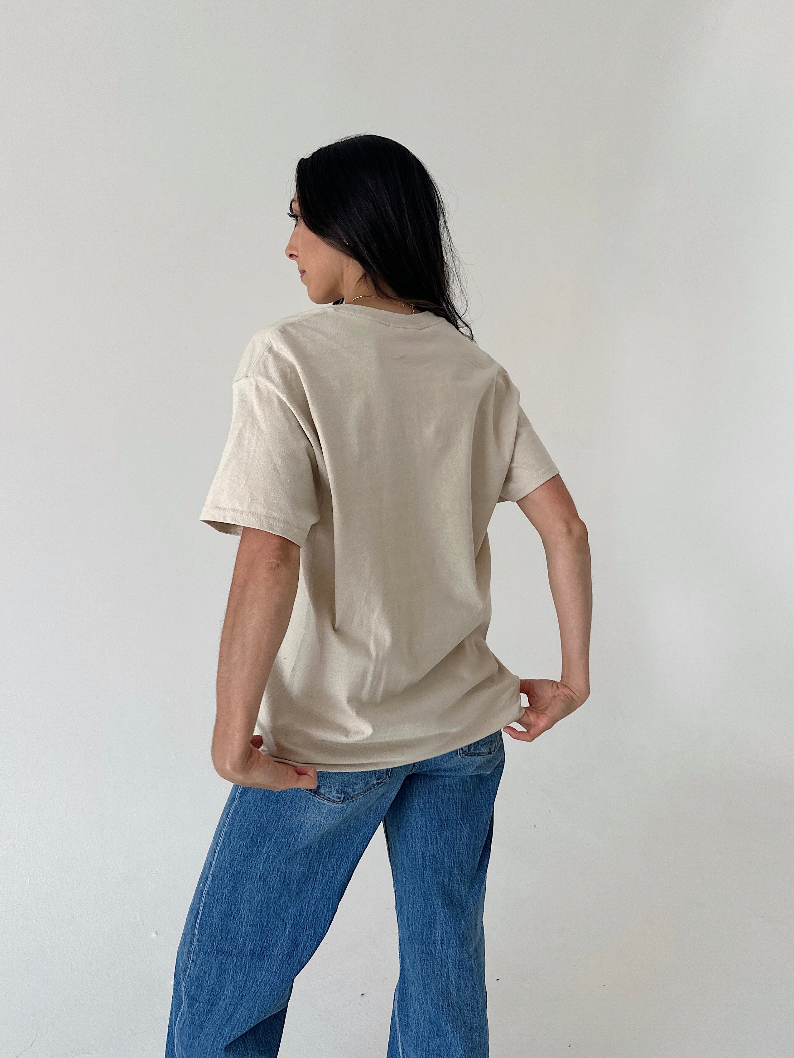 A woman with long black hair is shown from behind wearing an oversized beige t-shirt and blue jeans, standing against a plain off-white wall.