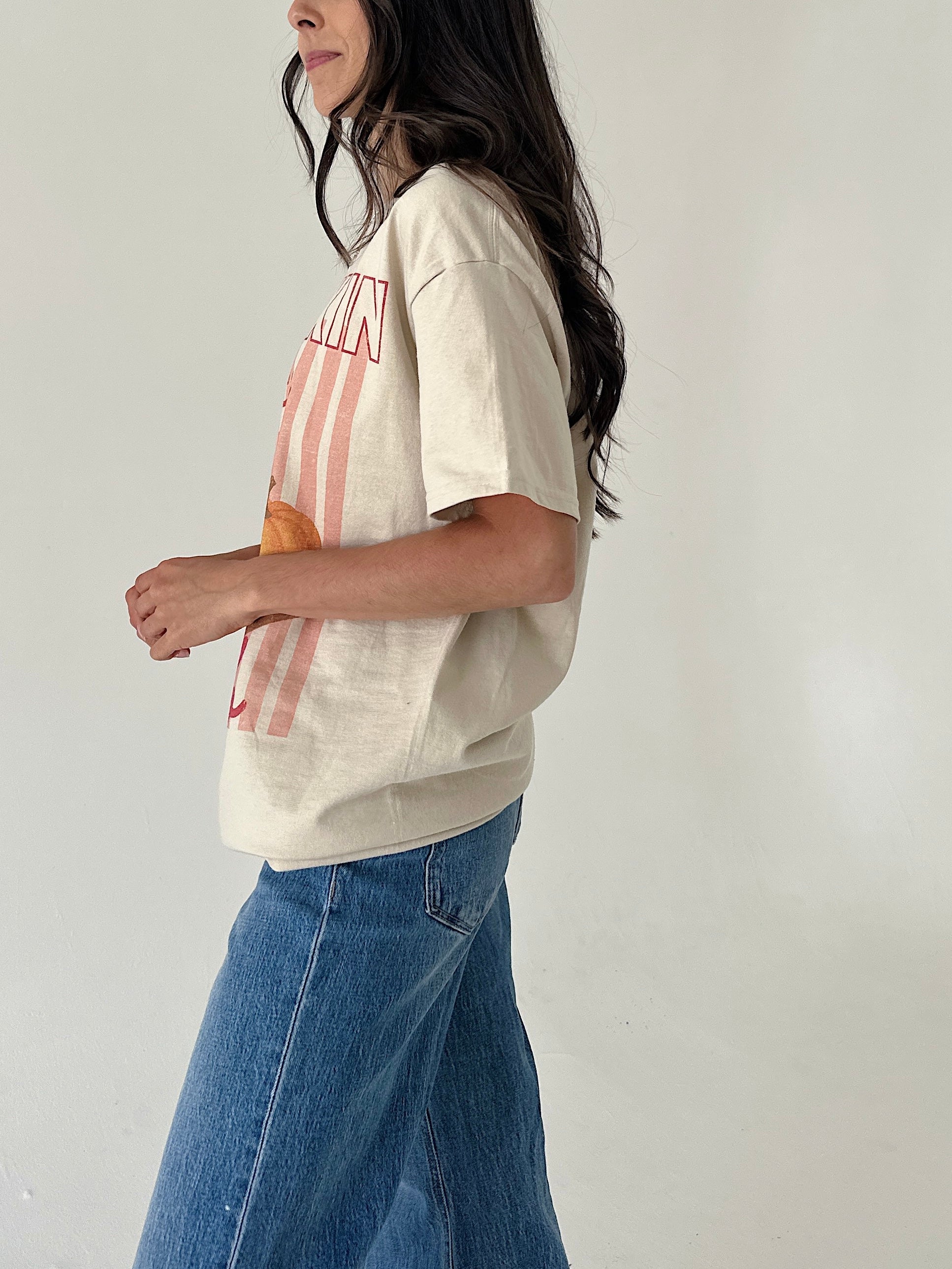 A woman wearing a cream-colored graphic t-shirt with pink stripes and blue denim jeans, shown from a side profile against a plain off-white wall.