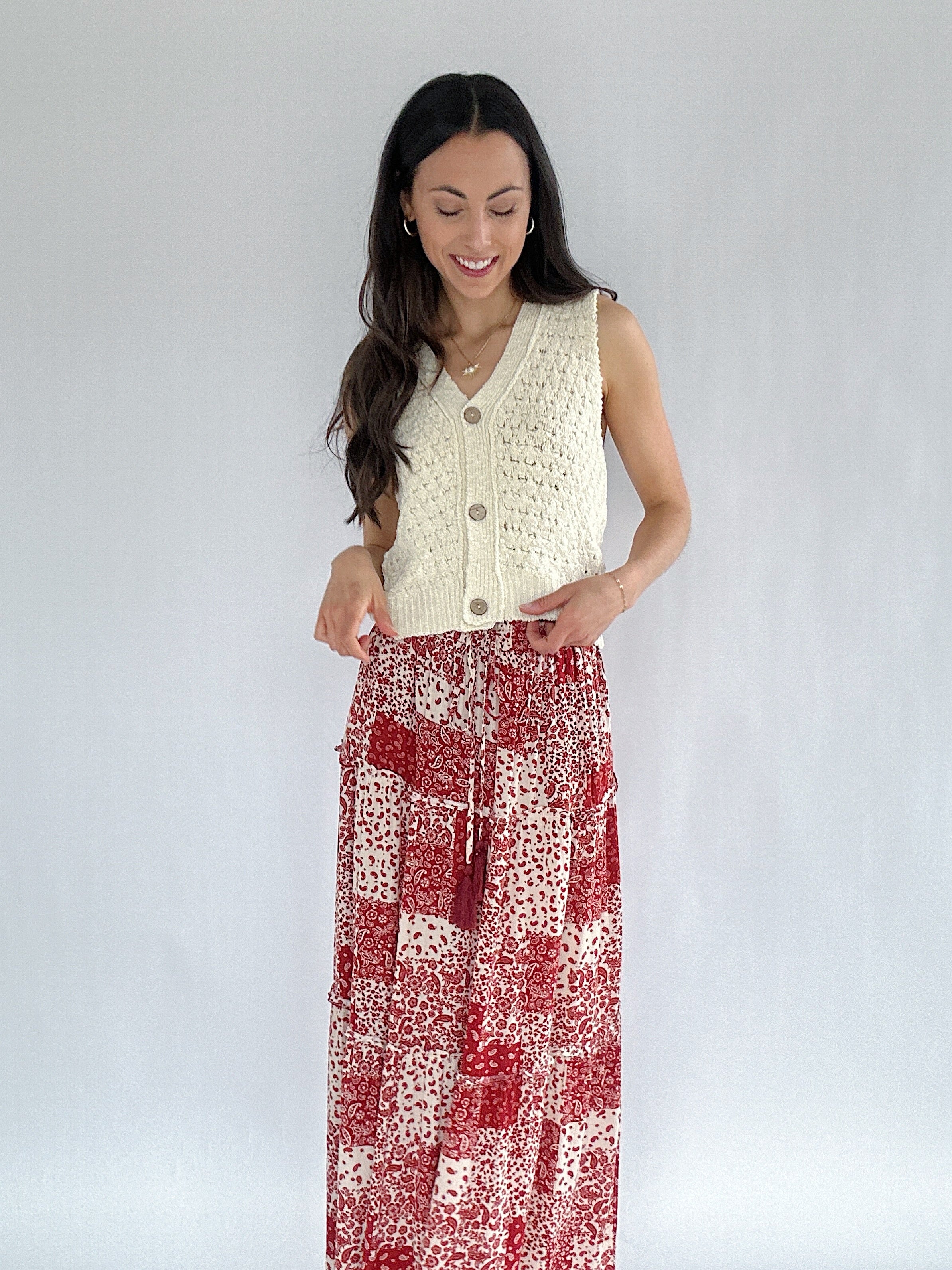 Woman wearing a cream sleeveless knitted vest over a red and white patterned dress on a plain background
