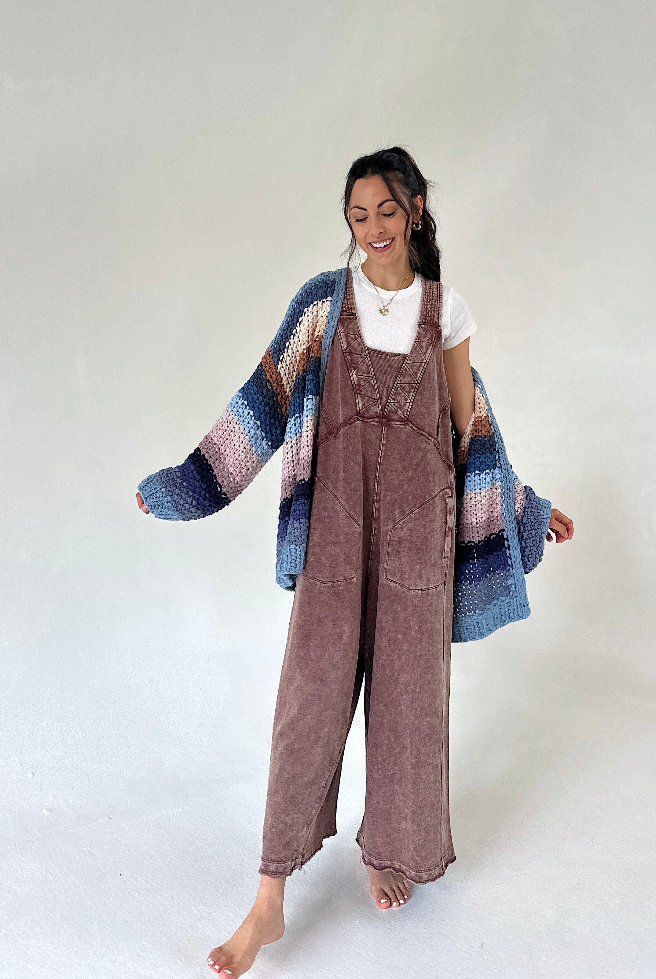 A woman stands barefoot against a plain light background wearing a brown terry knit overall jumpsuit, a white t-shirt, and a chunky knit cardigan with blue, pink, and purple stripes.