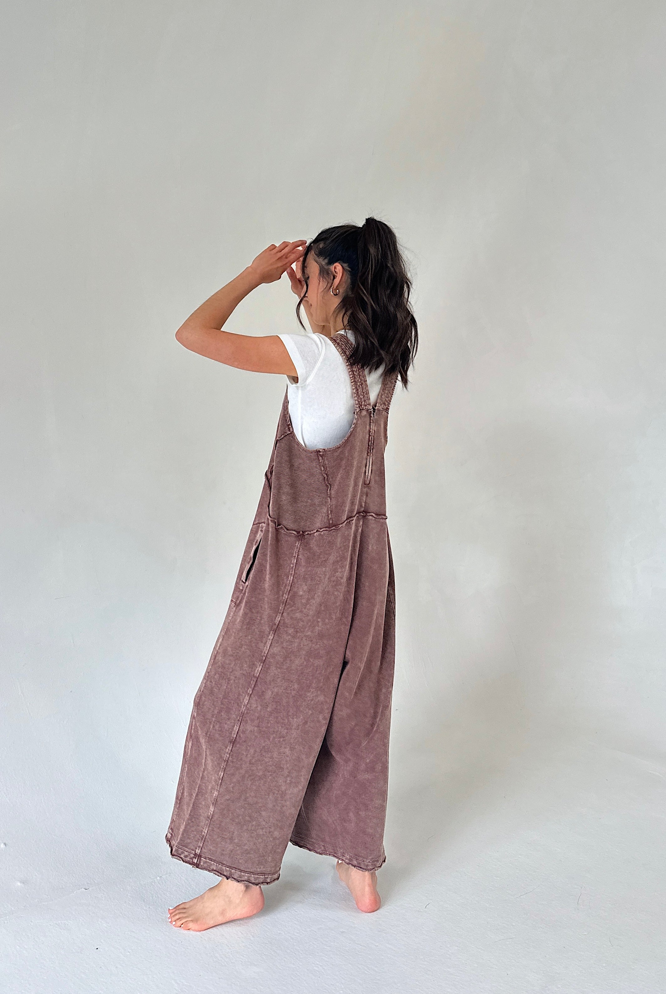 A woman stands barefoot against a plain light gray studio background, wearing a loose mauve-brown overall jumpsuit over a white short-sleeve top. She is shown from the back with her arms raised near her face.