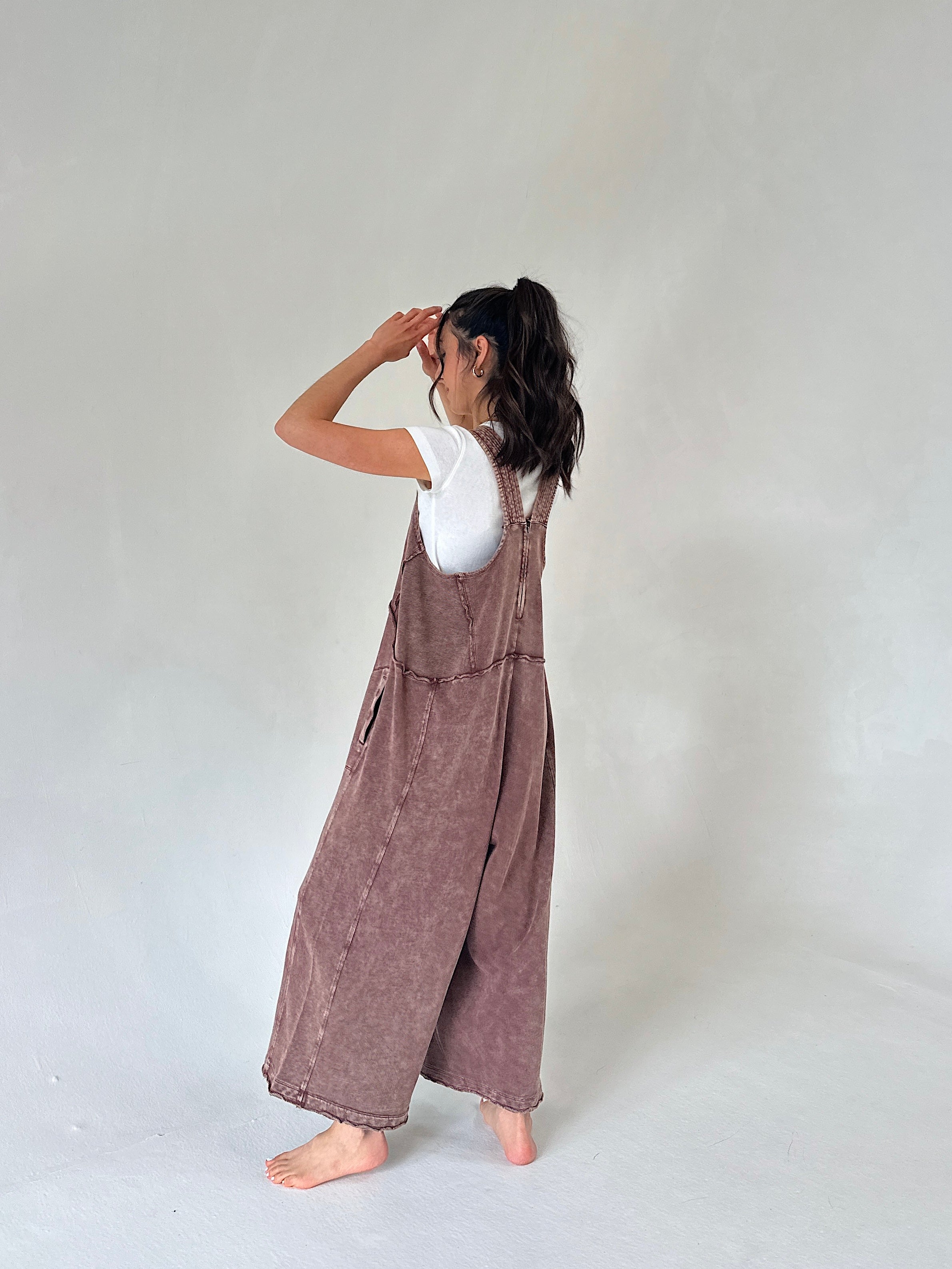 A woman stands barefoot against a plain light gray studio background, wearing a loose mauve-brown overall jumpsuit over a white short-sleeve top. She is shown from the back with her arms raised near her face.