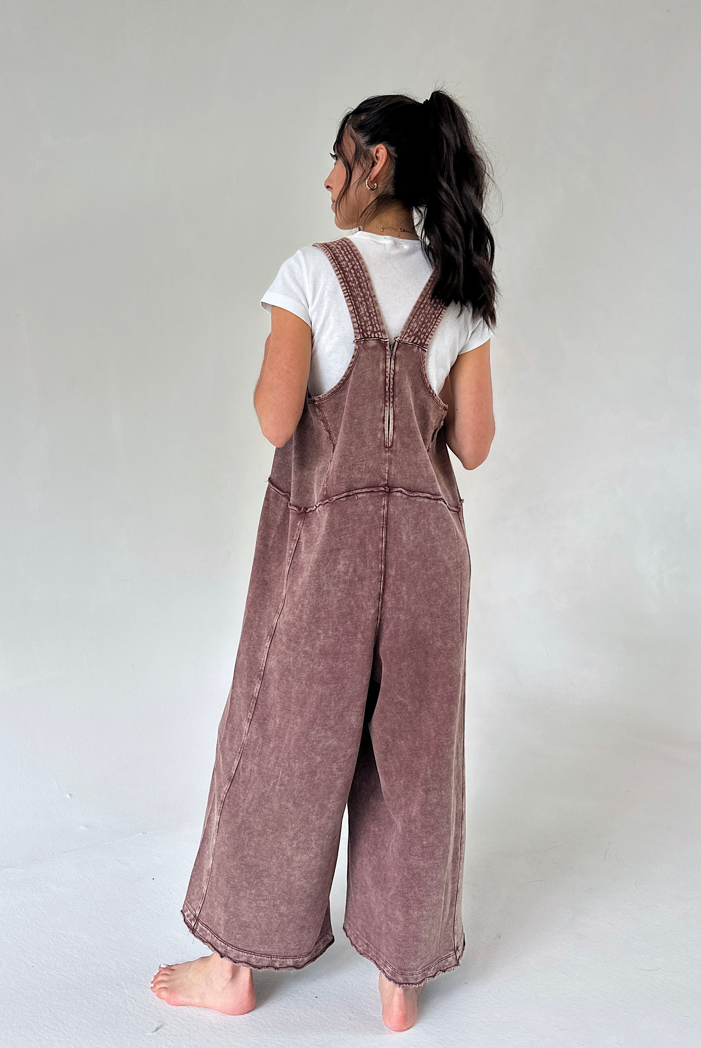 A model shown from the back wearing a dusty mauve-brown wide-leg terry overall jumpsuit over a white t-shirt, standing barefoot against a neutral light gray studio background.