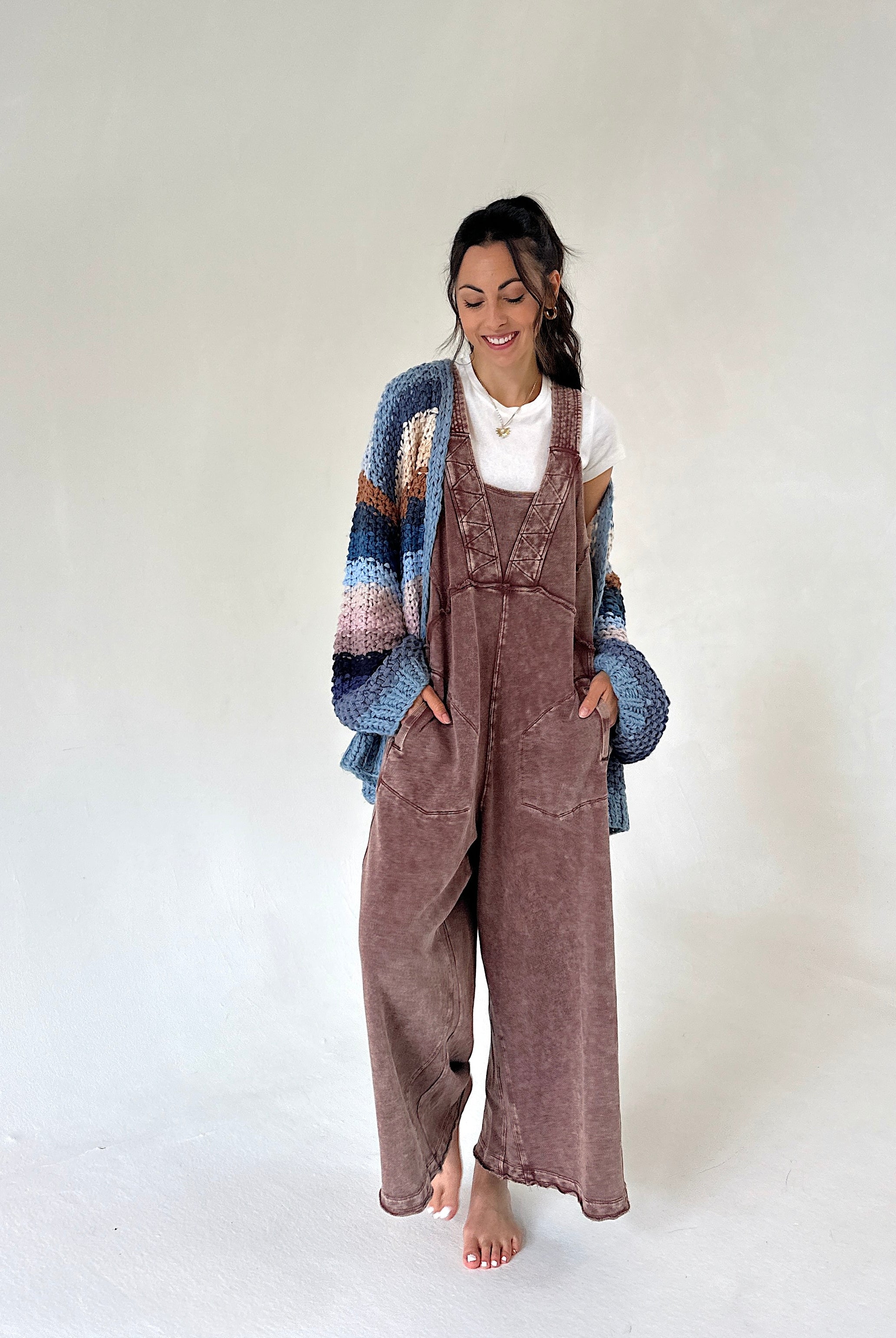 A woman stands barefoot against a plain light gray studio background, wearing a brown terry overall jumpsuit with a white top and a chunky knit cardigan in blue, beige, and brown tones.