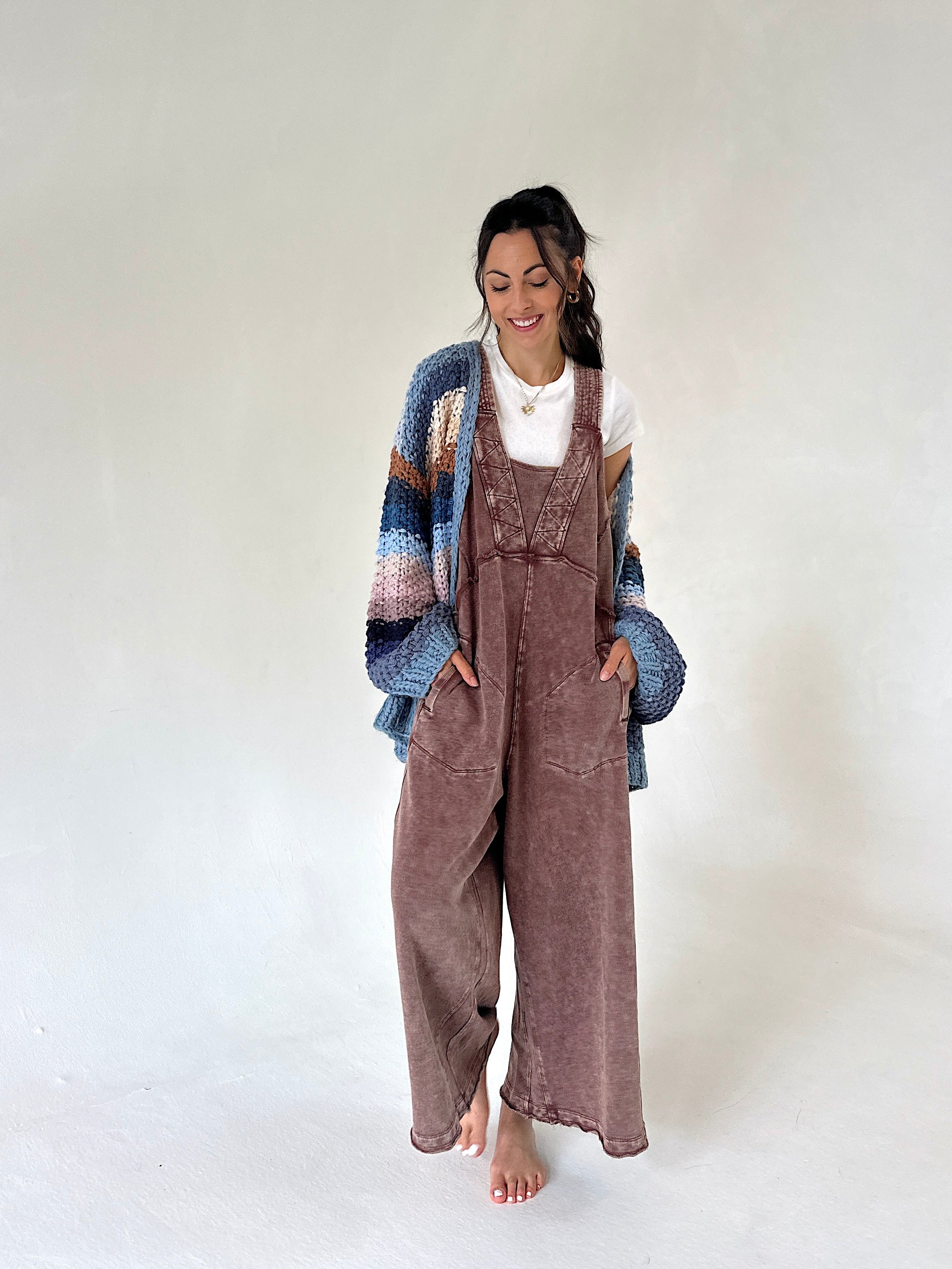 A woman stands barefoot against a plain light gray studio background, wearing a brown terry overall jumpsuit with a white top and a chunky knit cardigan in blue, beige, and brown tones.
