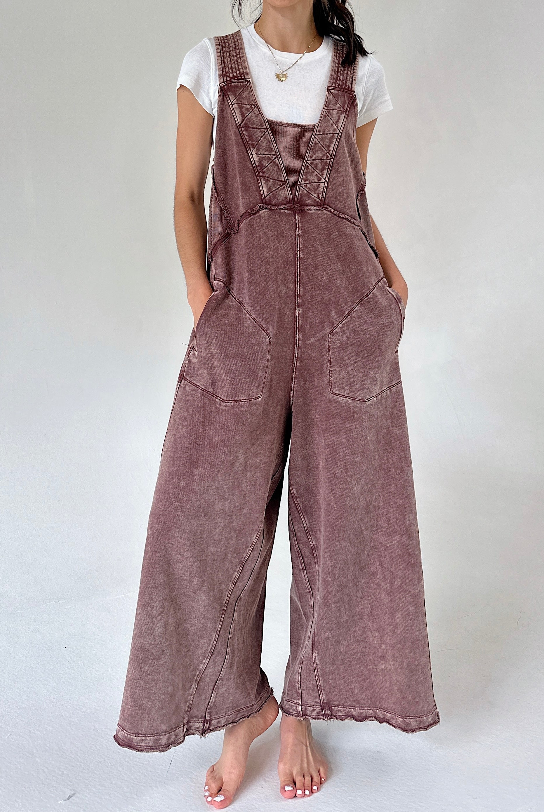 A woman wearing a brownish mauve wide-leg terry overall jumpsuit over a white t-shirt, standing against a light gray studio background.
