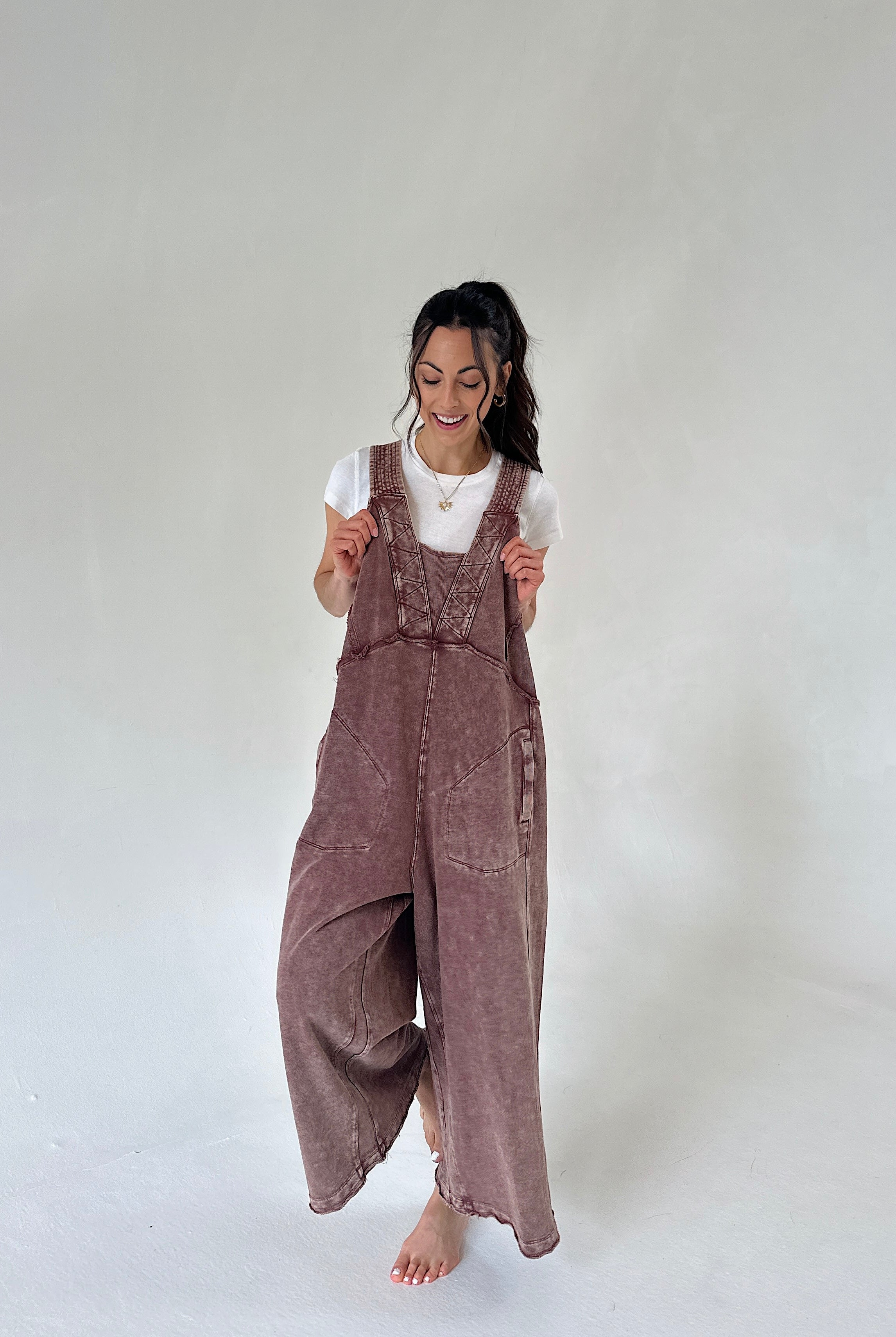 A woman wearing a dusty mauve-brown terry knit overall jumpsuit over a white t-shirt, standing barefoot against a neutral light gray studio background.