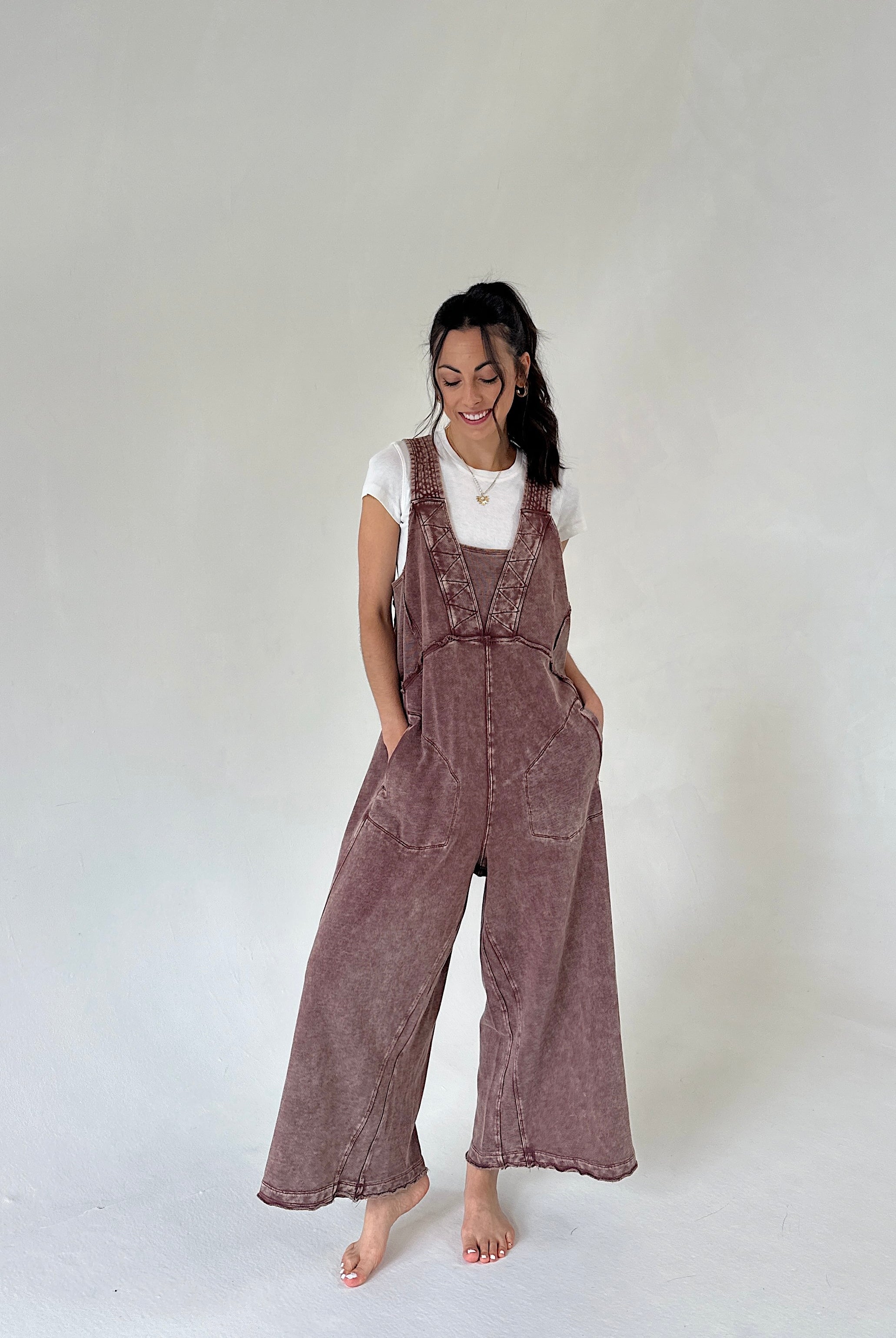 A woman modeling a brownish mauve wide-leg terry jumpsuit over a white t-shirt, standing against a neutral light gray studio background.