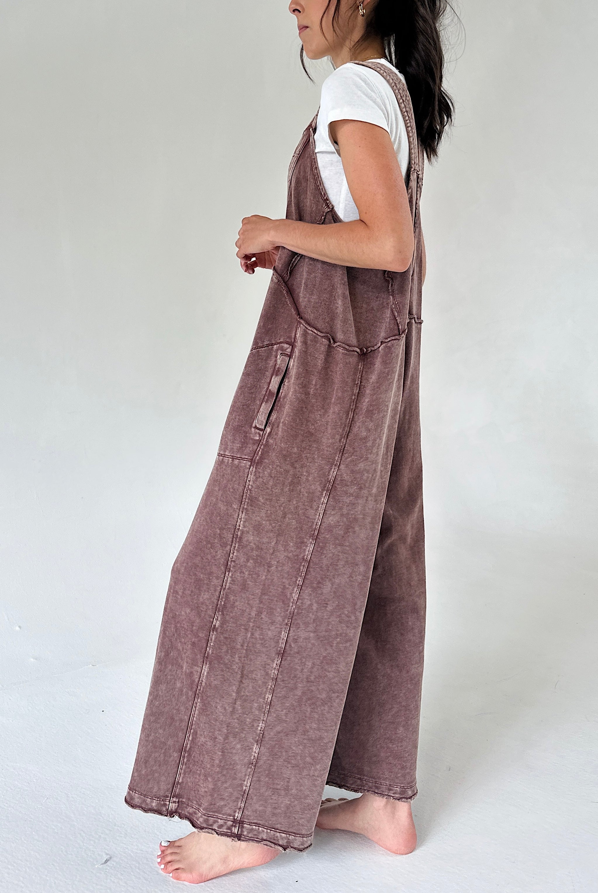 A woman shown from the side wearing a dusty brownish mauve wide-leg terry knit overall jumpsuit over a white t-shirt, standing barefoot against a plain light gray studio background.