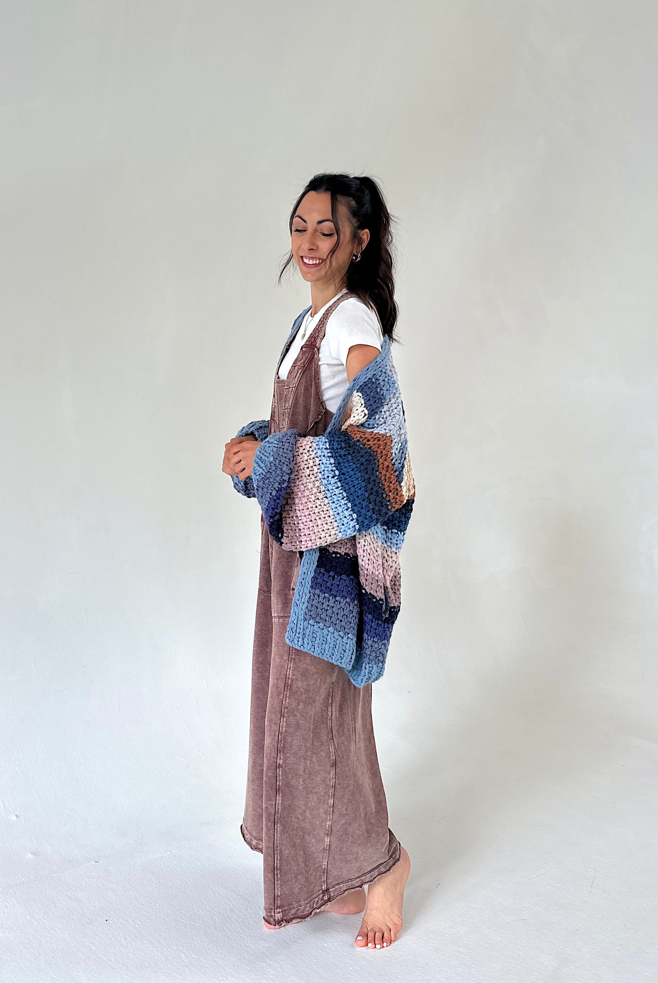 A woman stands barefoot against a plain light gray studio background, wearing a long mauve-brown jumpsuit over a white top and holding a chunky knit patchwork throw in blue, pink, and brown tones.