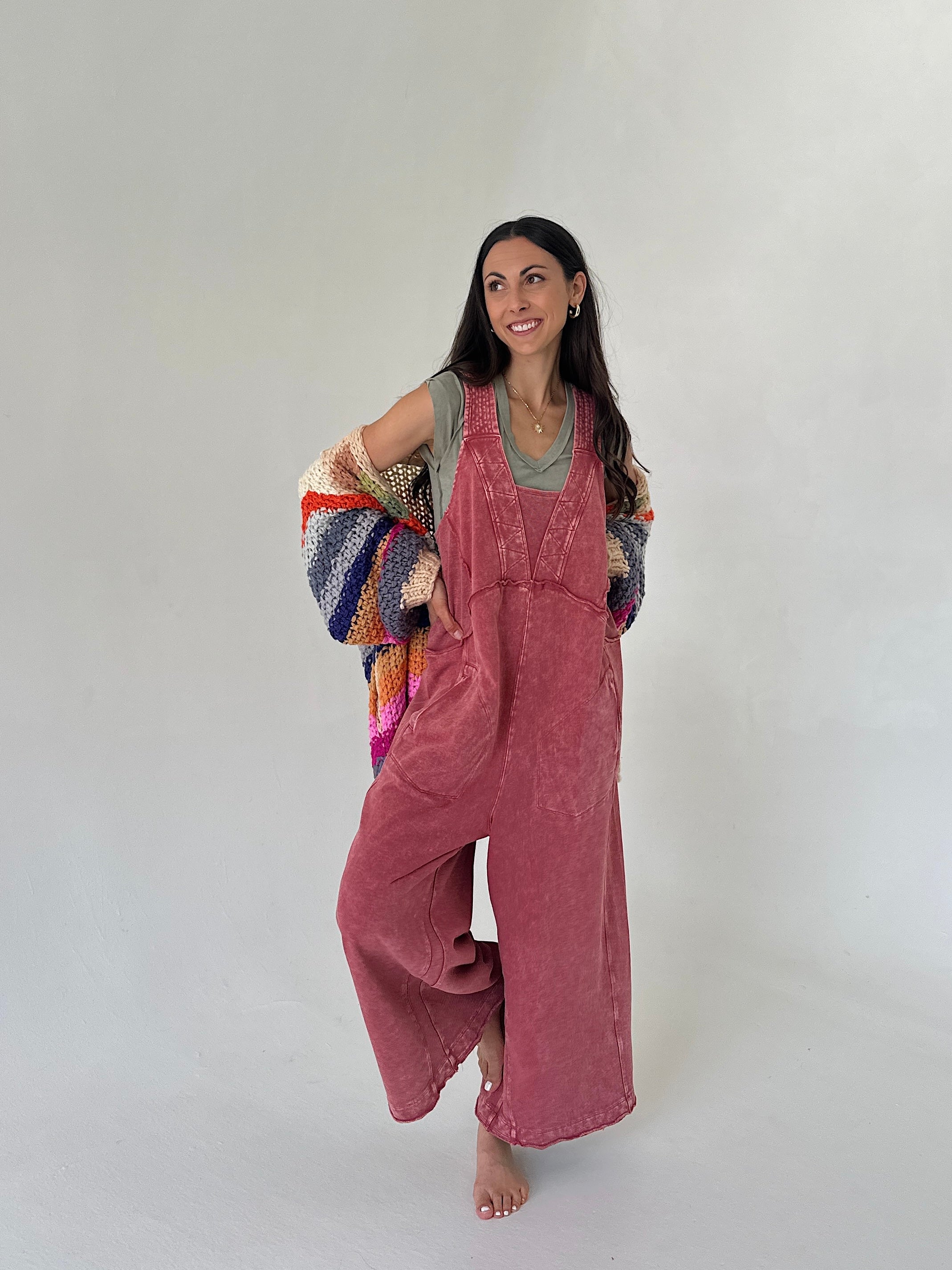 Woman wearing pink terry knit overalls against a white background with a colorful knit cardigan on arms