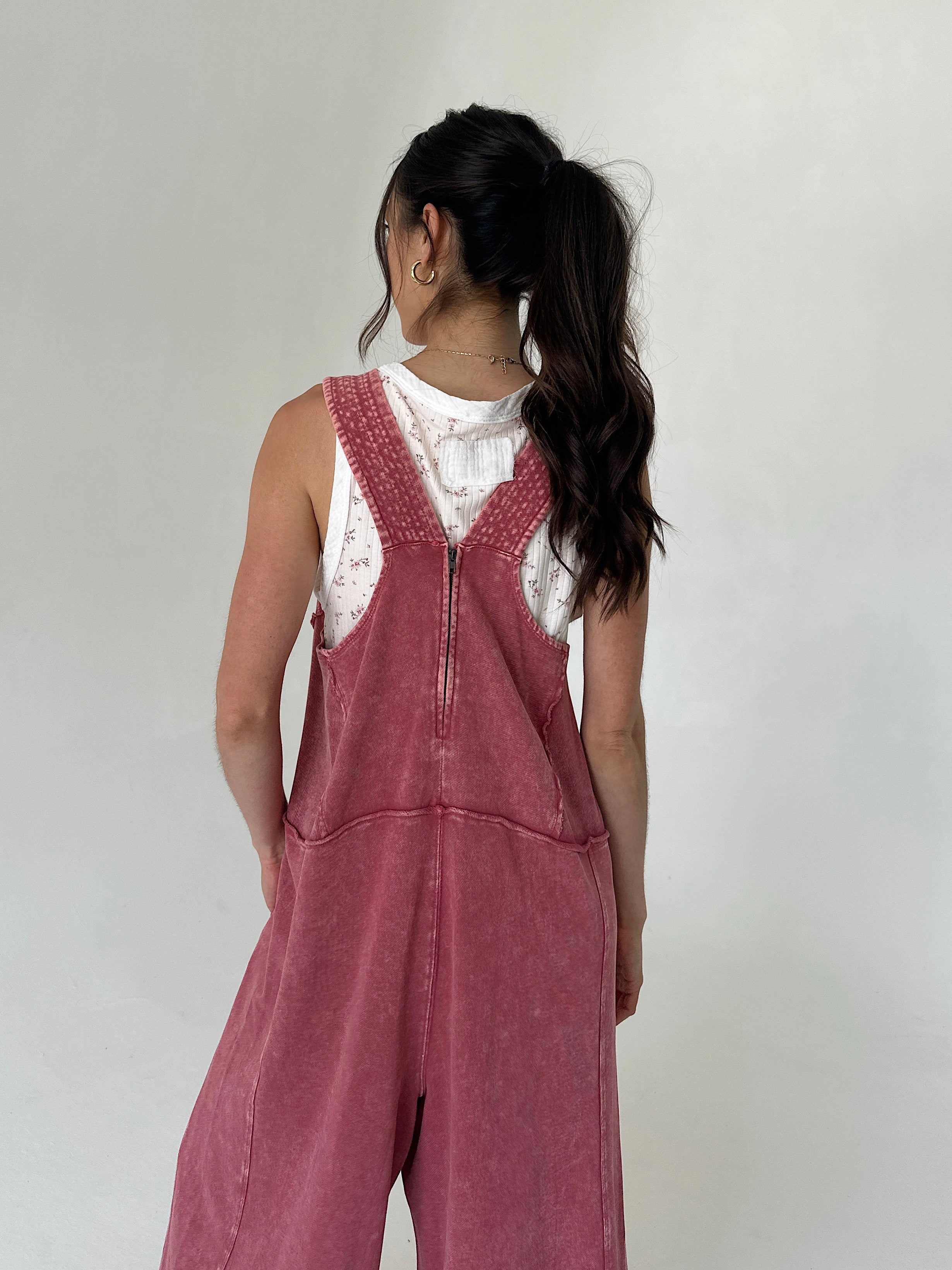 Person wearing a pink terry overalls with a white tank top against a plain background showing back zipper detail