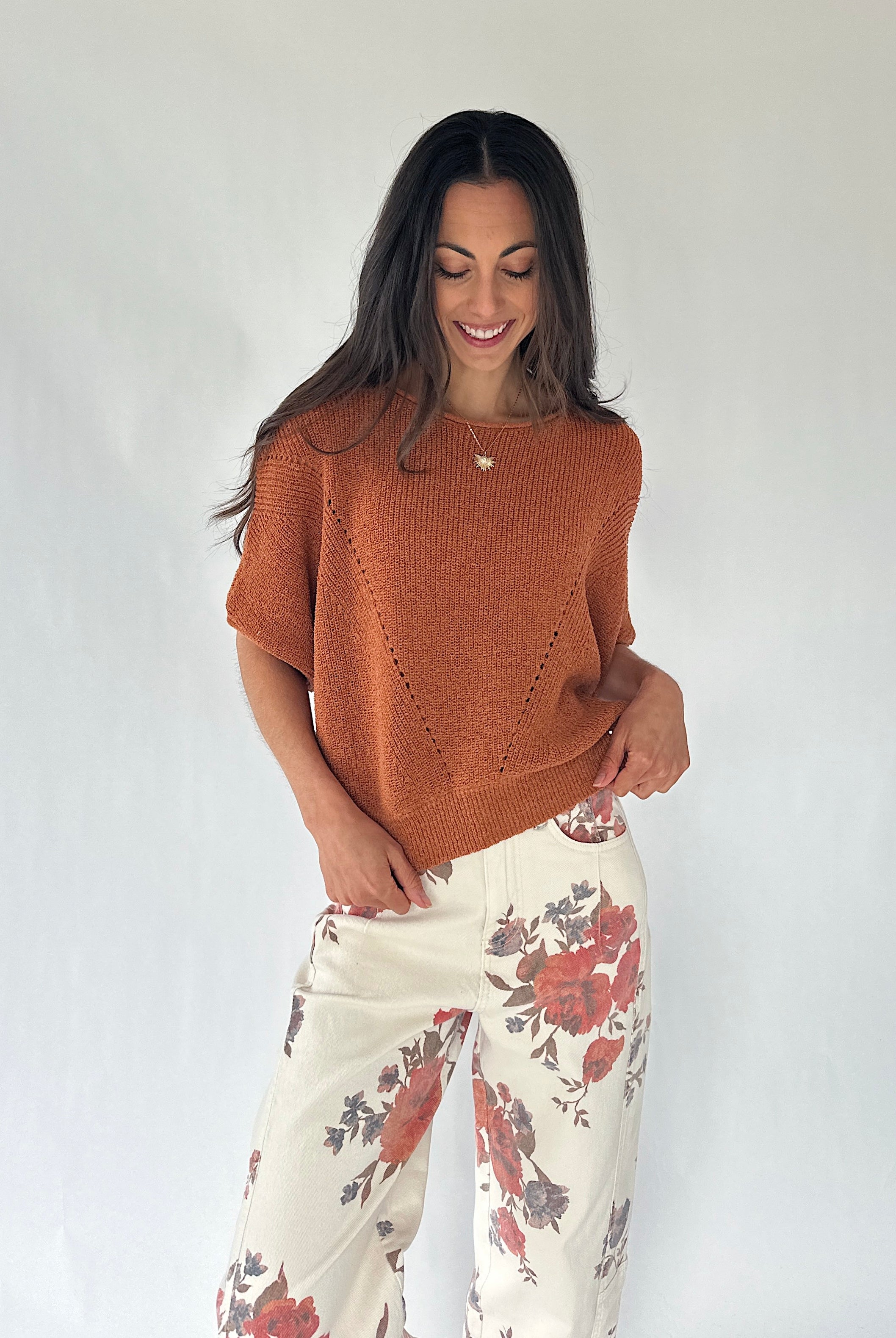 A woman modeling a short-sleeve knit top in a warm rust orange color, paired with cream wide-leg pants featuring a red and gray floral pattern, against a plain white studio background.