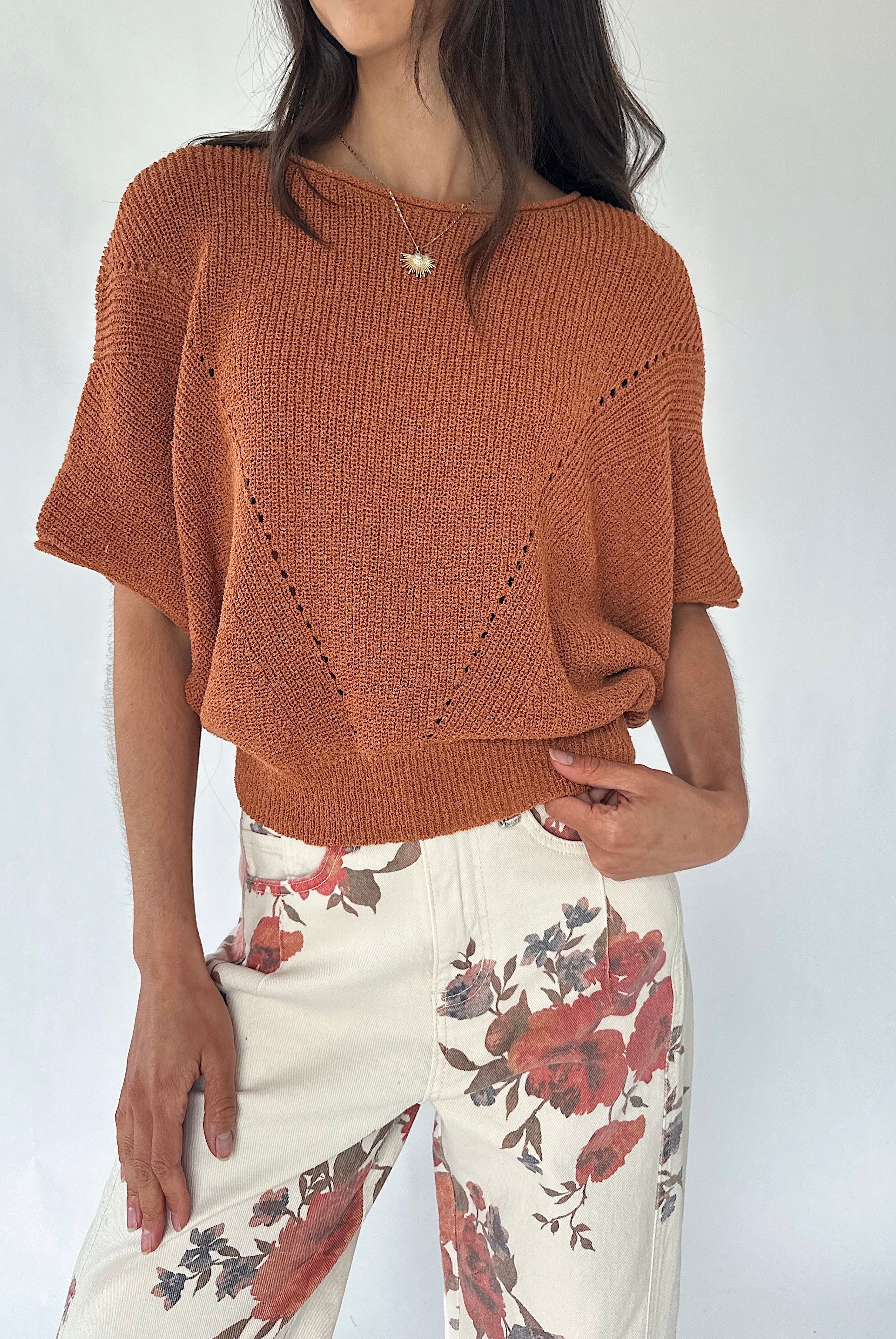 A woman wearing a rust-orange knit short-sleeve sweater with textured openwork details and cream floral print pants, photographed against a plain light background.