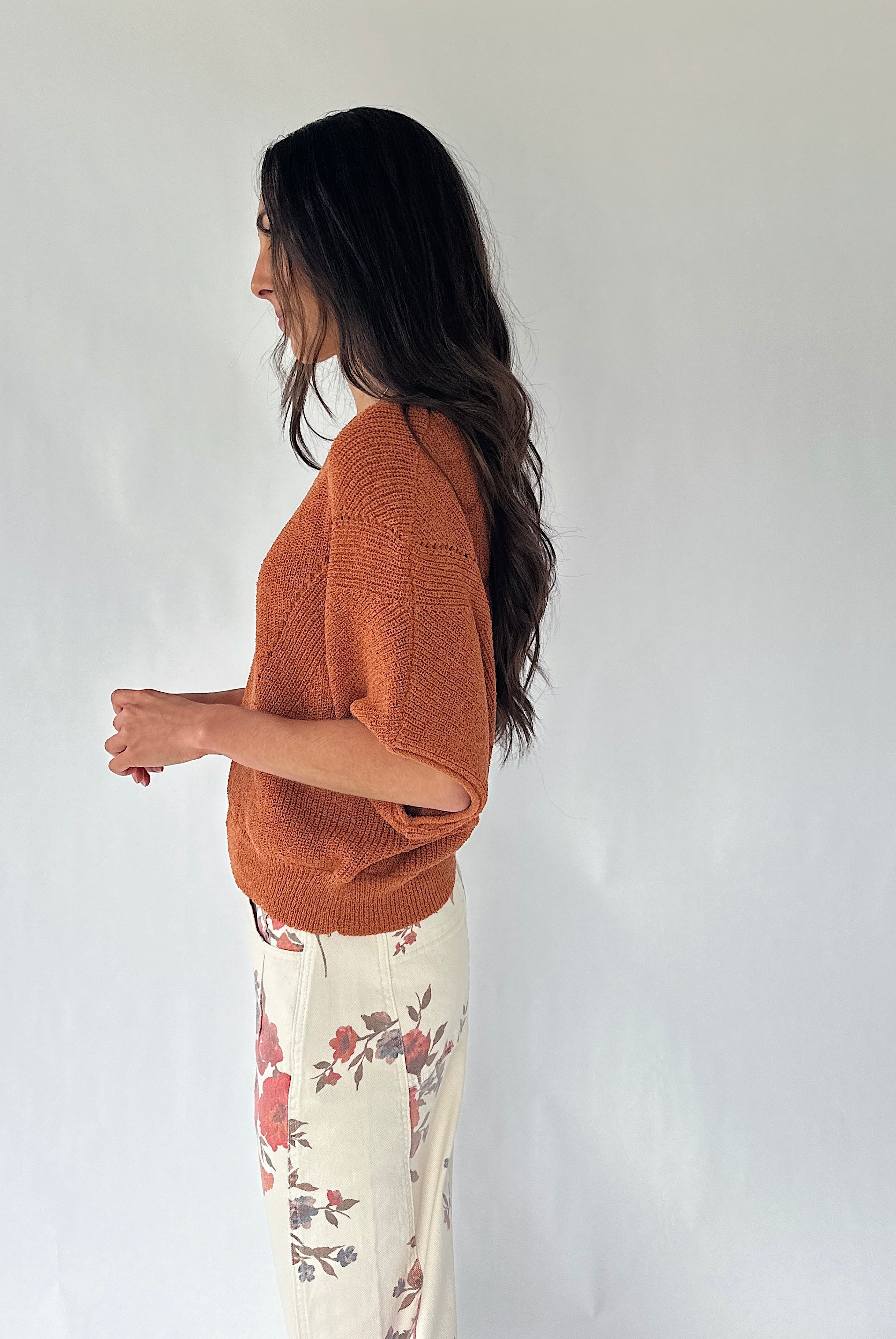 A woman shown in side profile wearing a textured rust-orange knit sweater and cream floral pants against a plain light gray wall.
