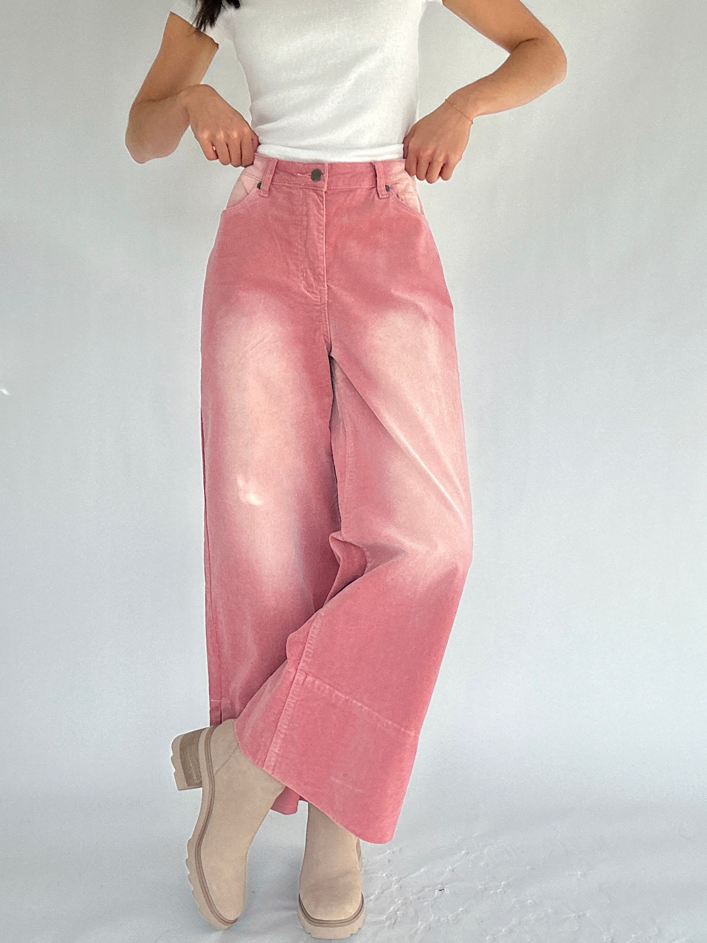 Person wearing pink corduroy pants and a white top on a plain background with legs crossed