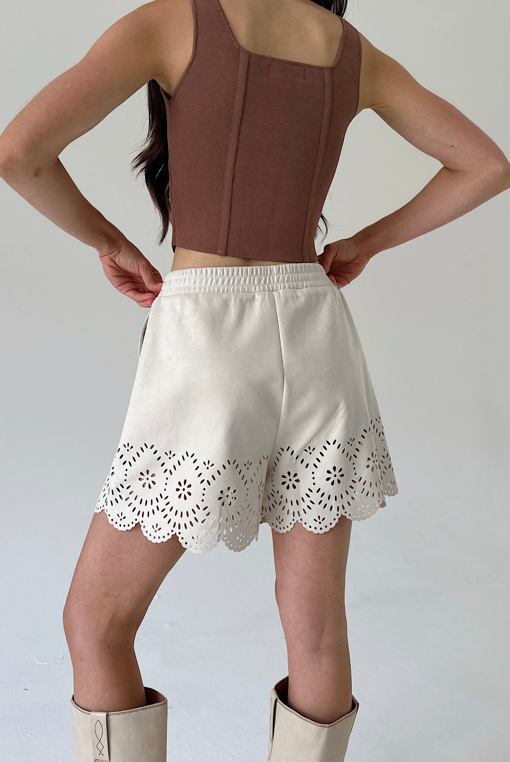 A model shown from the back wearing a brown sleeveless crop top and cream-colored shorts with a scalloped eyelet lace hem, paired with cream boots against a plain light gray background.