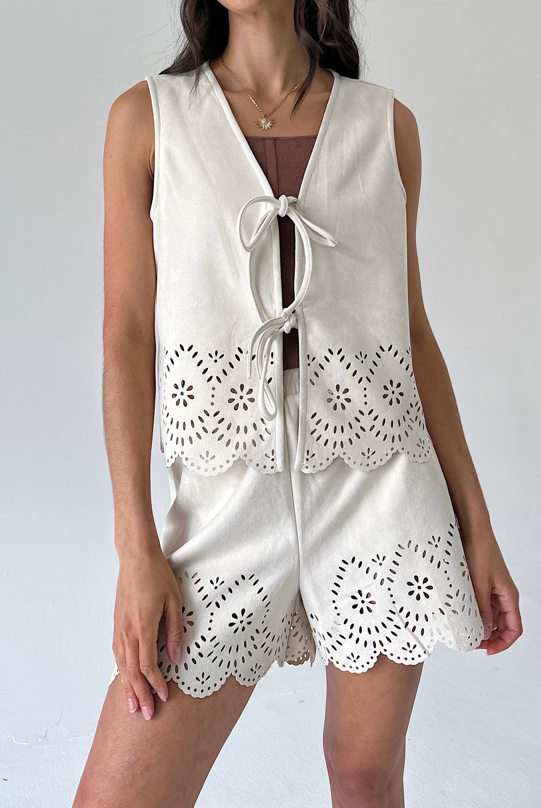 A woman wearing a sleeveless cream-colored vest with intricate eyelet lace detailing and a front tie closure, layered over a brown top with matching shorts