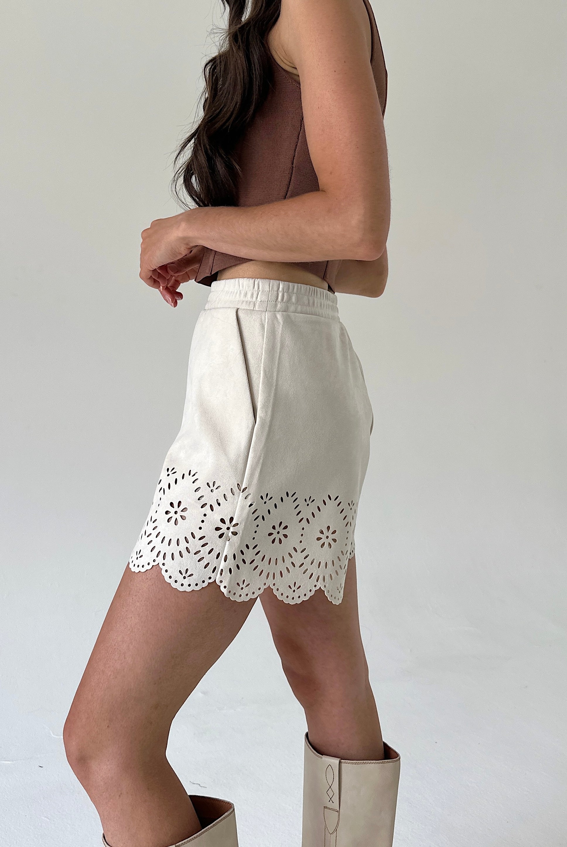 A model is shown from the side wearing off-white eyelet shorts with a scalloped floral cutwork hem, paired with a brown sleeveless top and beige boots against a plain light gray studio background.