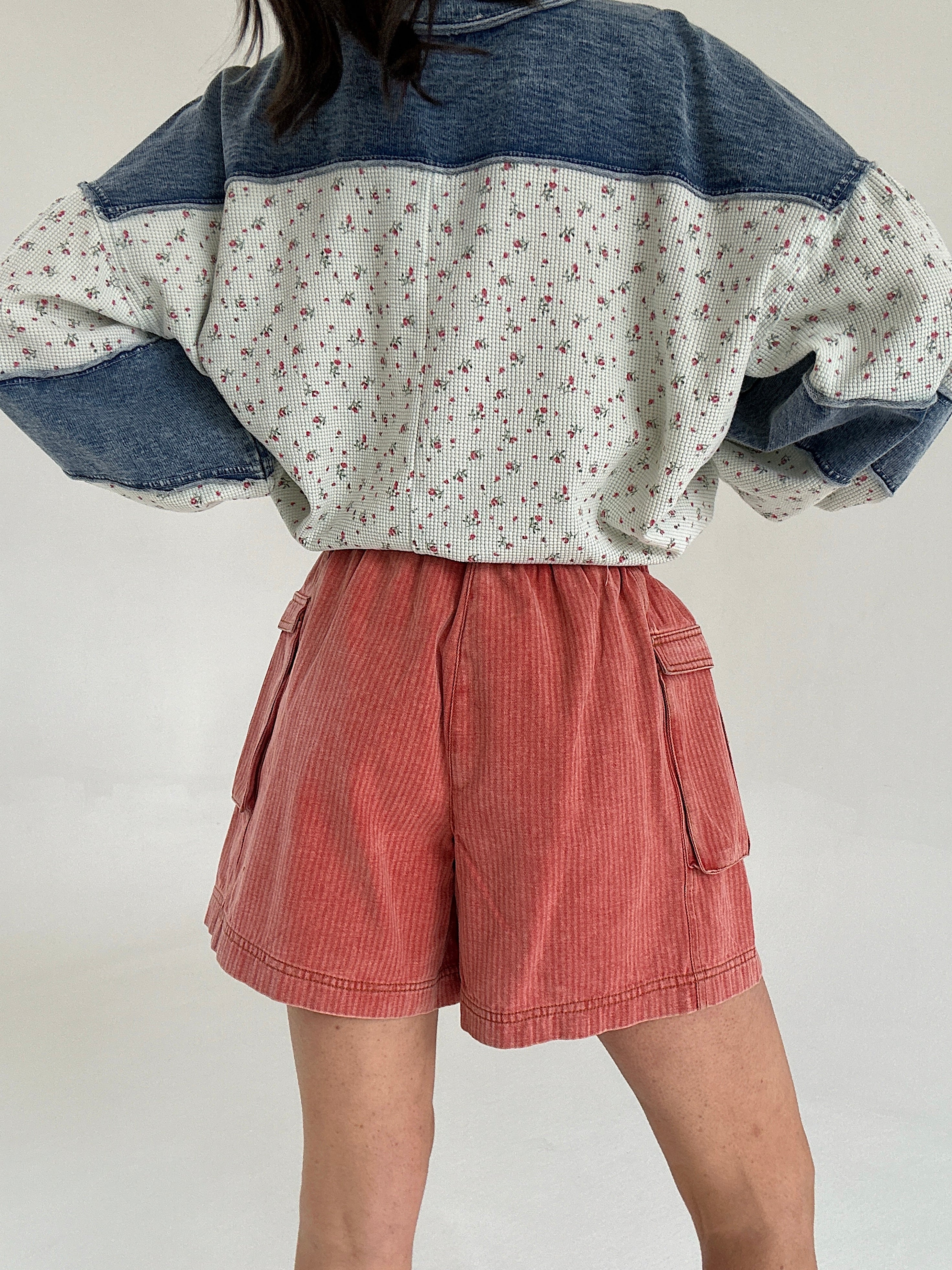 Person wearing a two-tone shirt with floral pattern and denim sleeves, paired with red cargo shorts on a plain background to show back