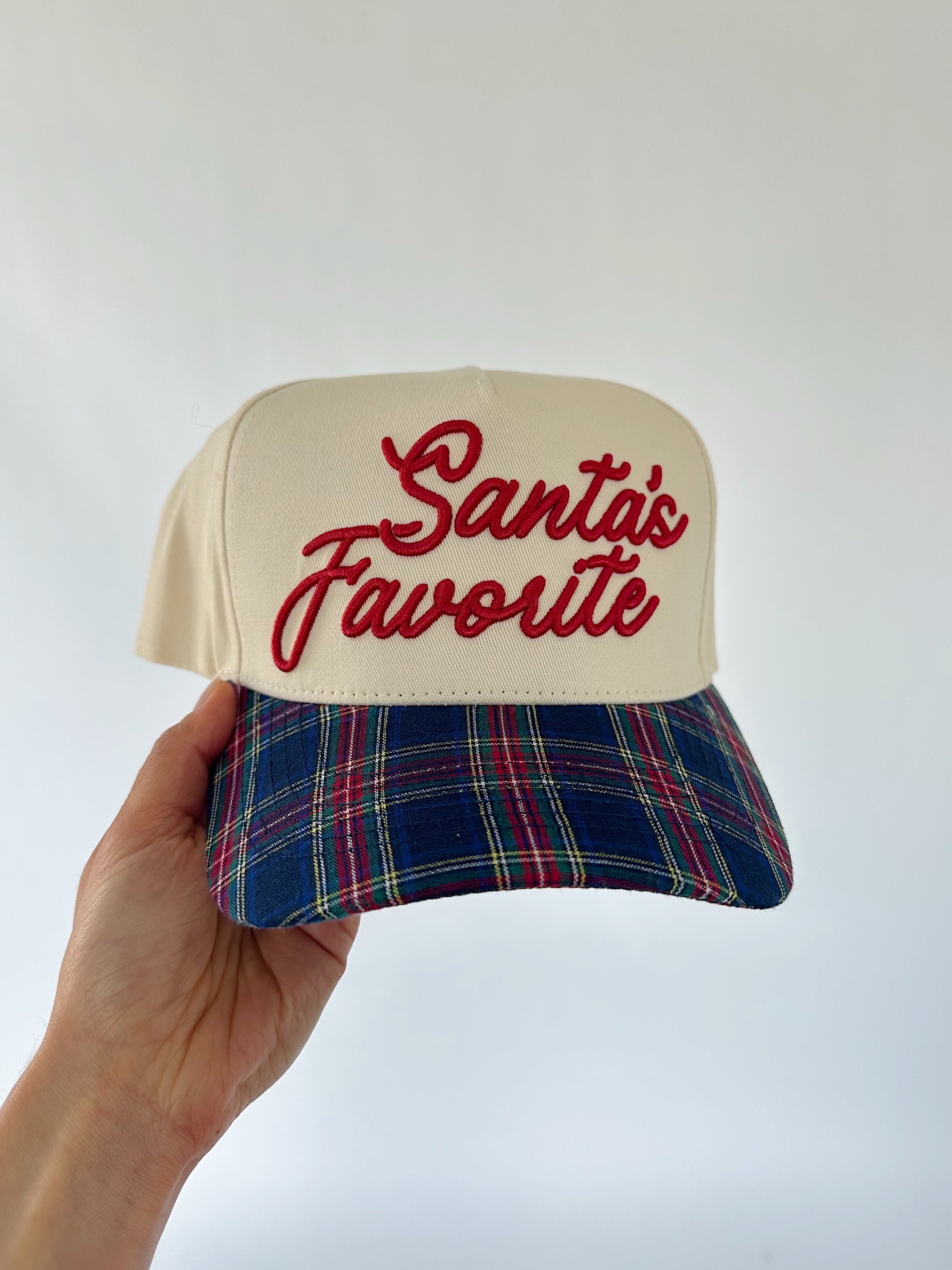 Cap with 'Santa's Favorite' text and plaid bill held by a hand against a white background