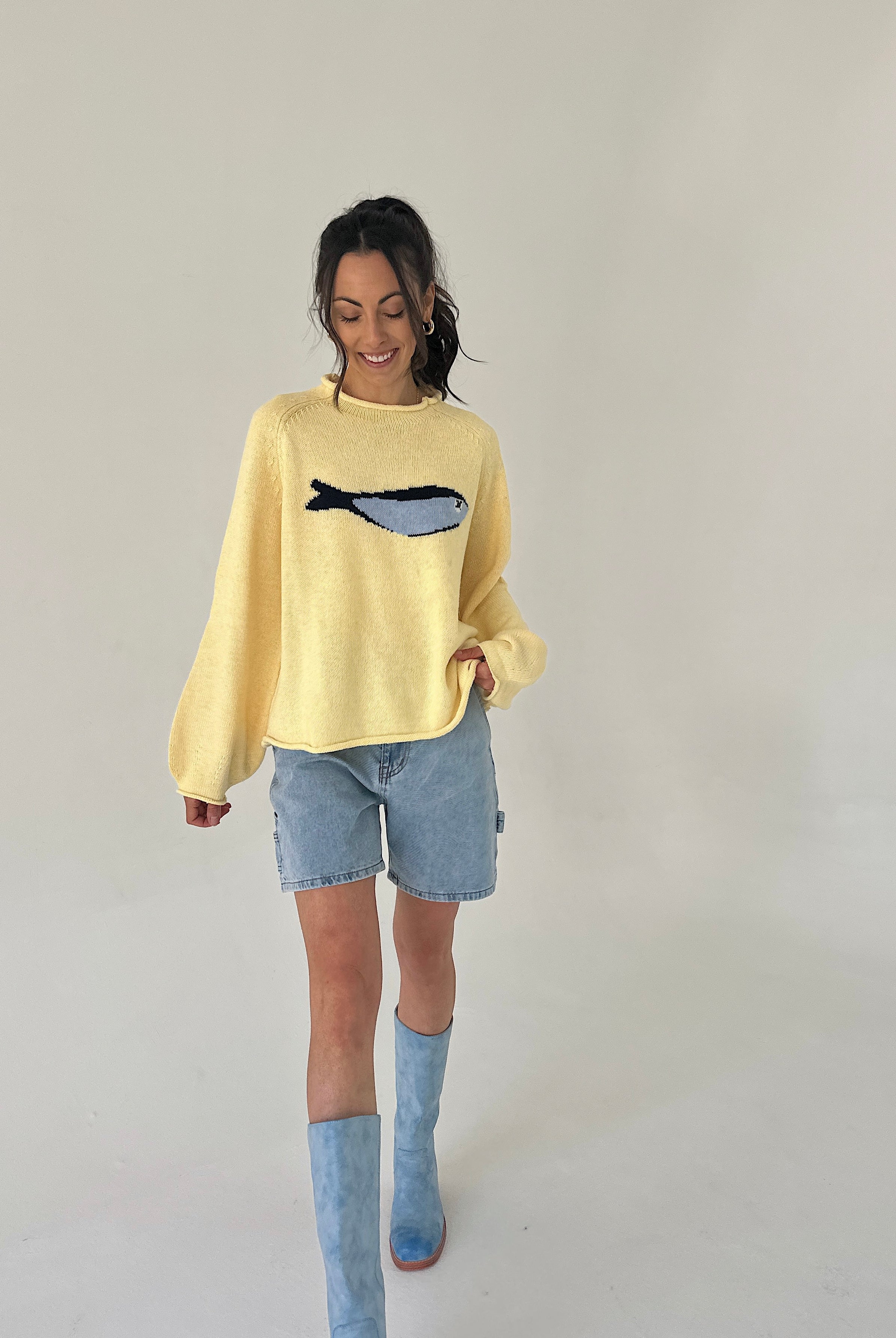 Woman wearing a yellow sweater with a fish design, denim shorts, and blue boots on a plain background