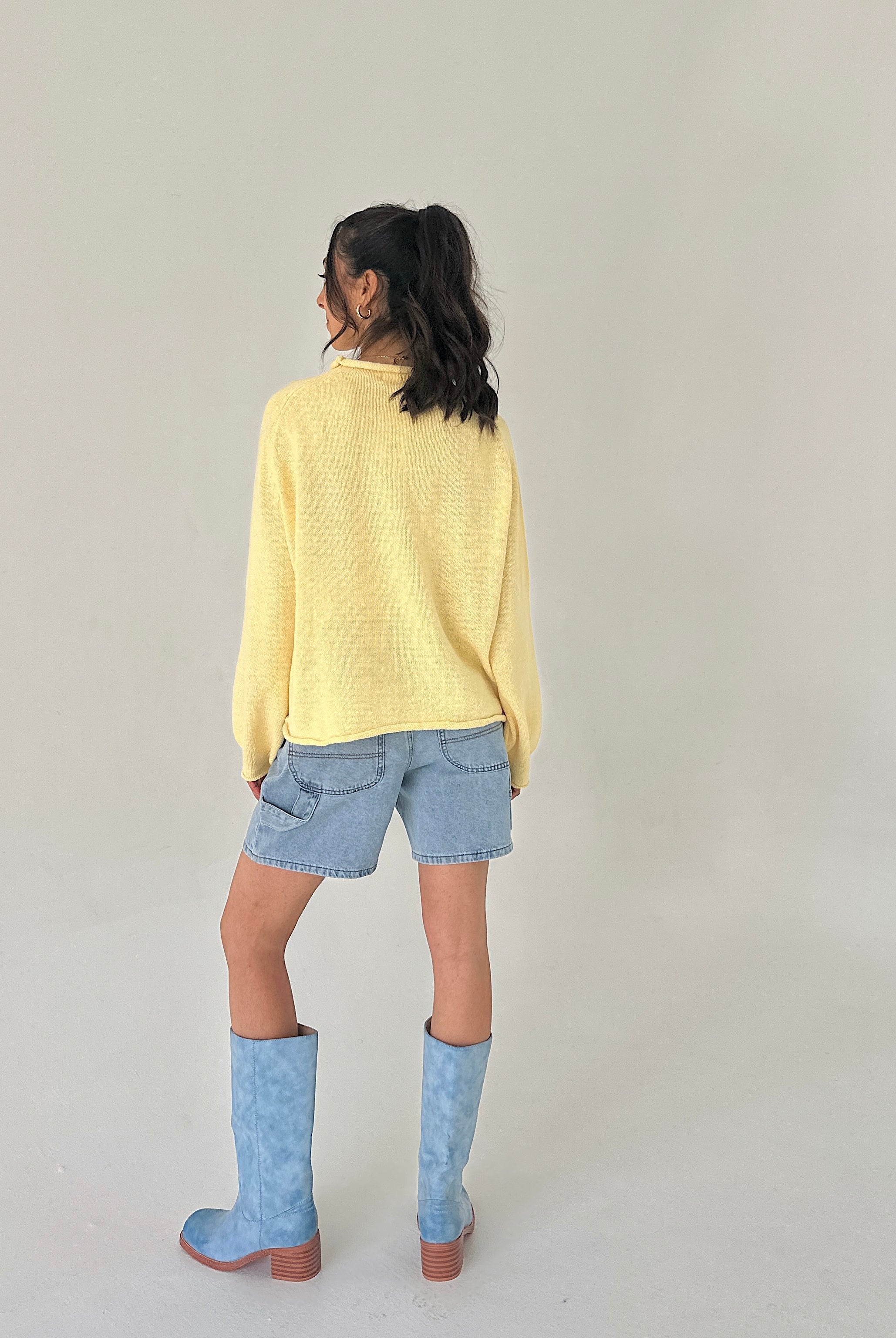 Person wearing a yellow sweater, denim shorts, and blue boots on a plain background from back