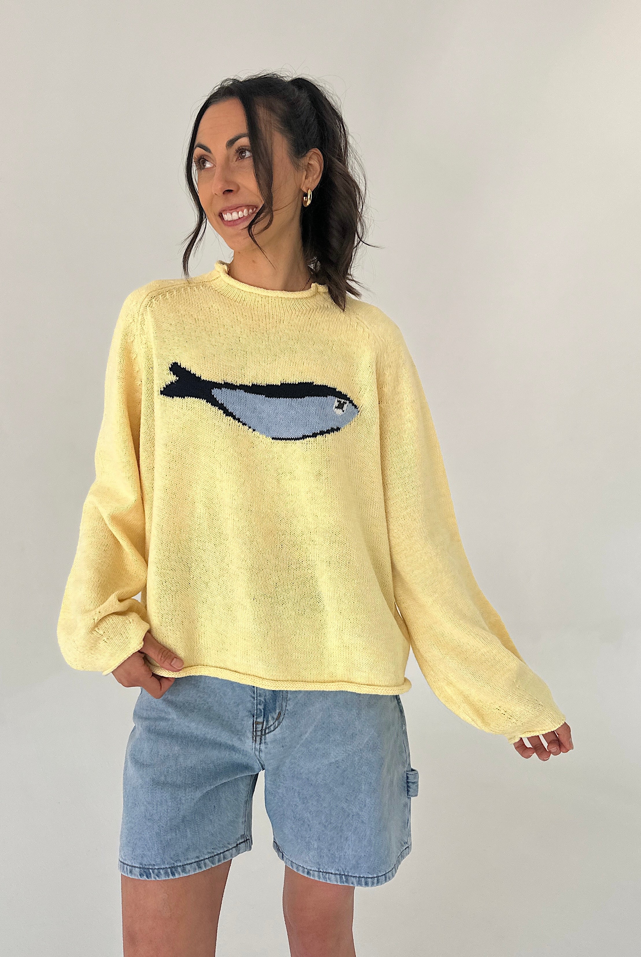 Woman wearing a yellow sweater with a fish design, standing against a plain background