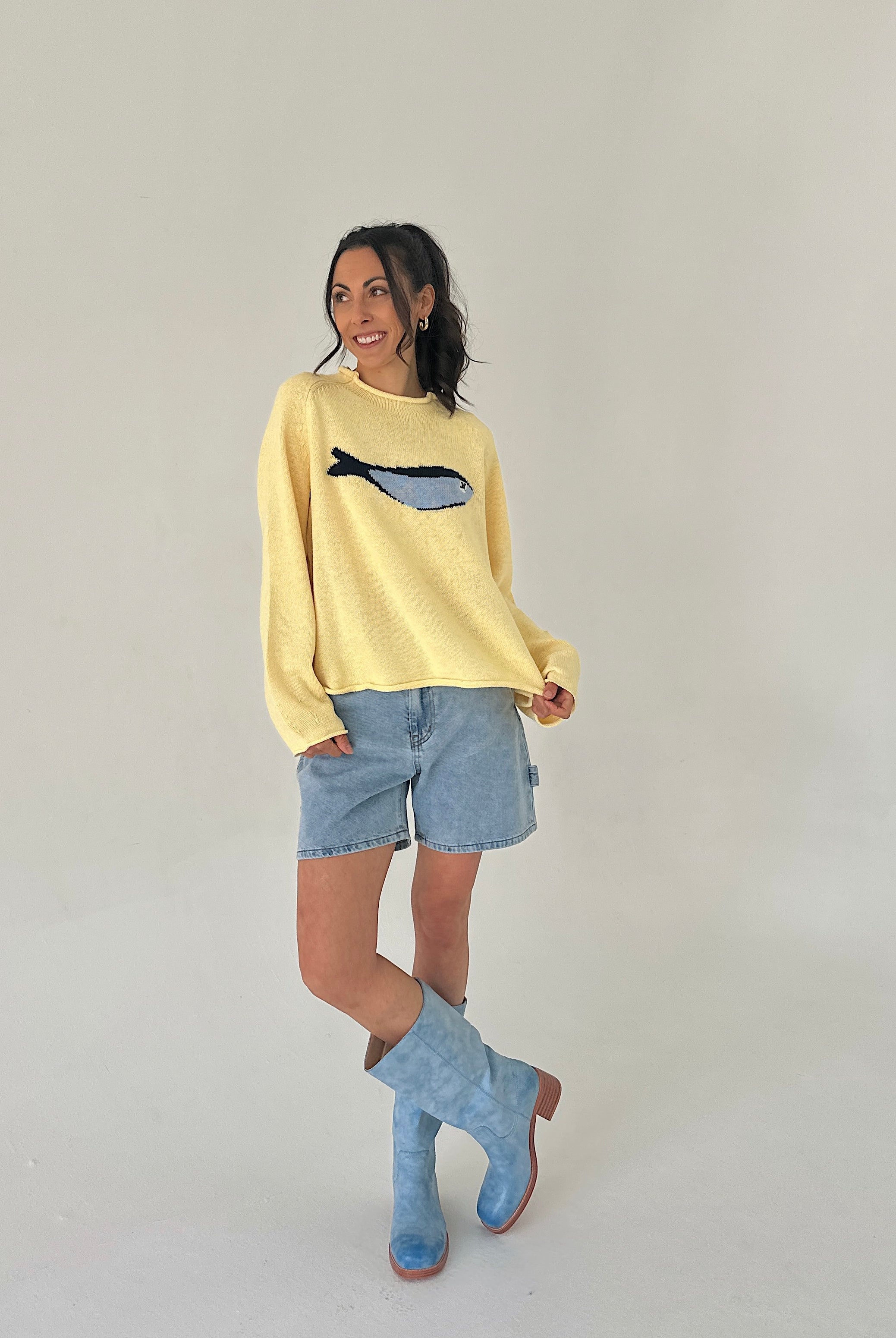 Woman wearing a yellow sweater with a fish design, denim shorts, and blue boots on a plain background
