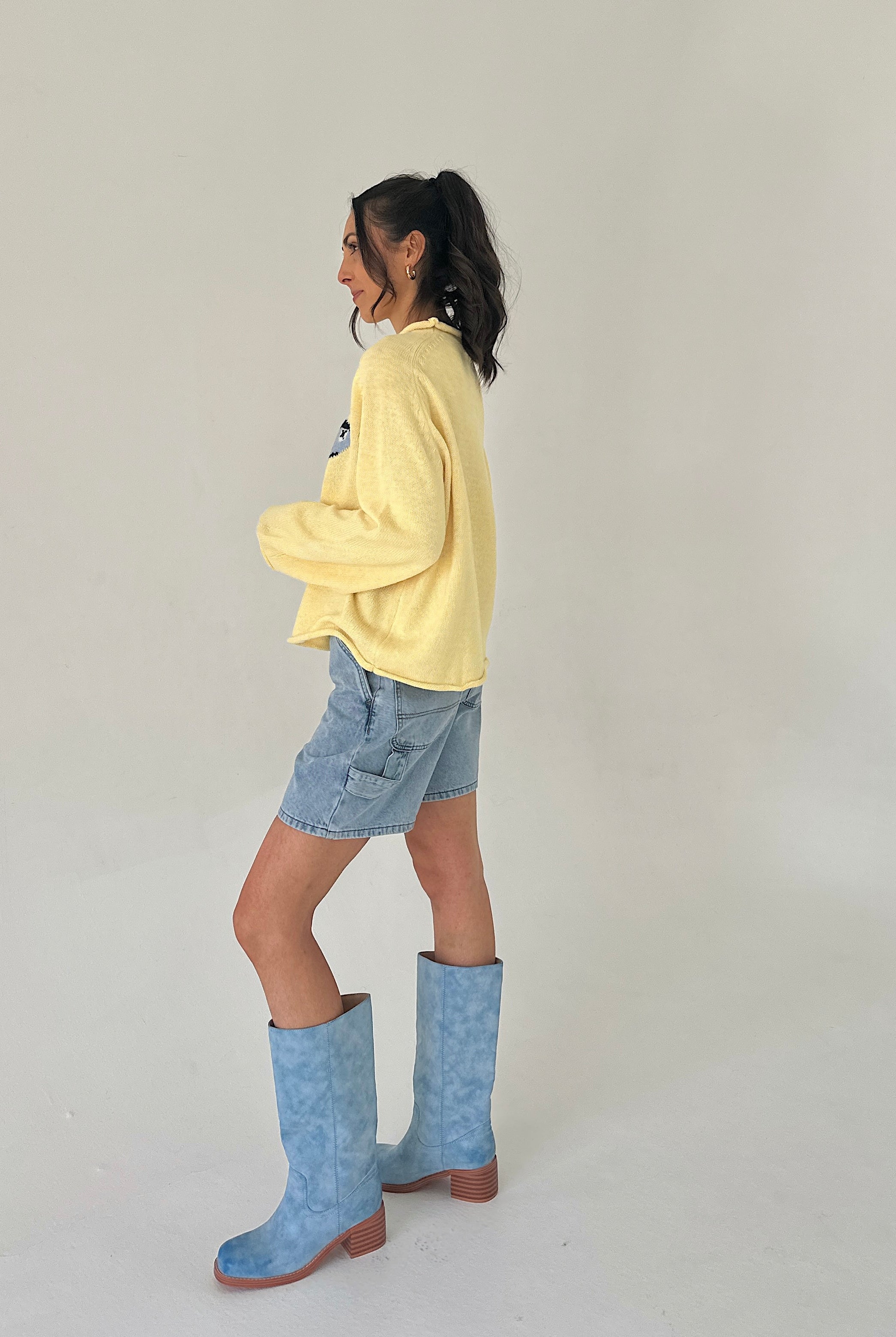 Person wearing a yellow sweater, denim shorts, and blue boots on a plain background