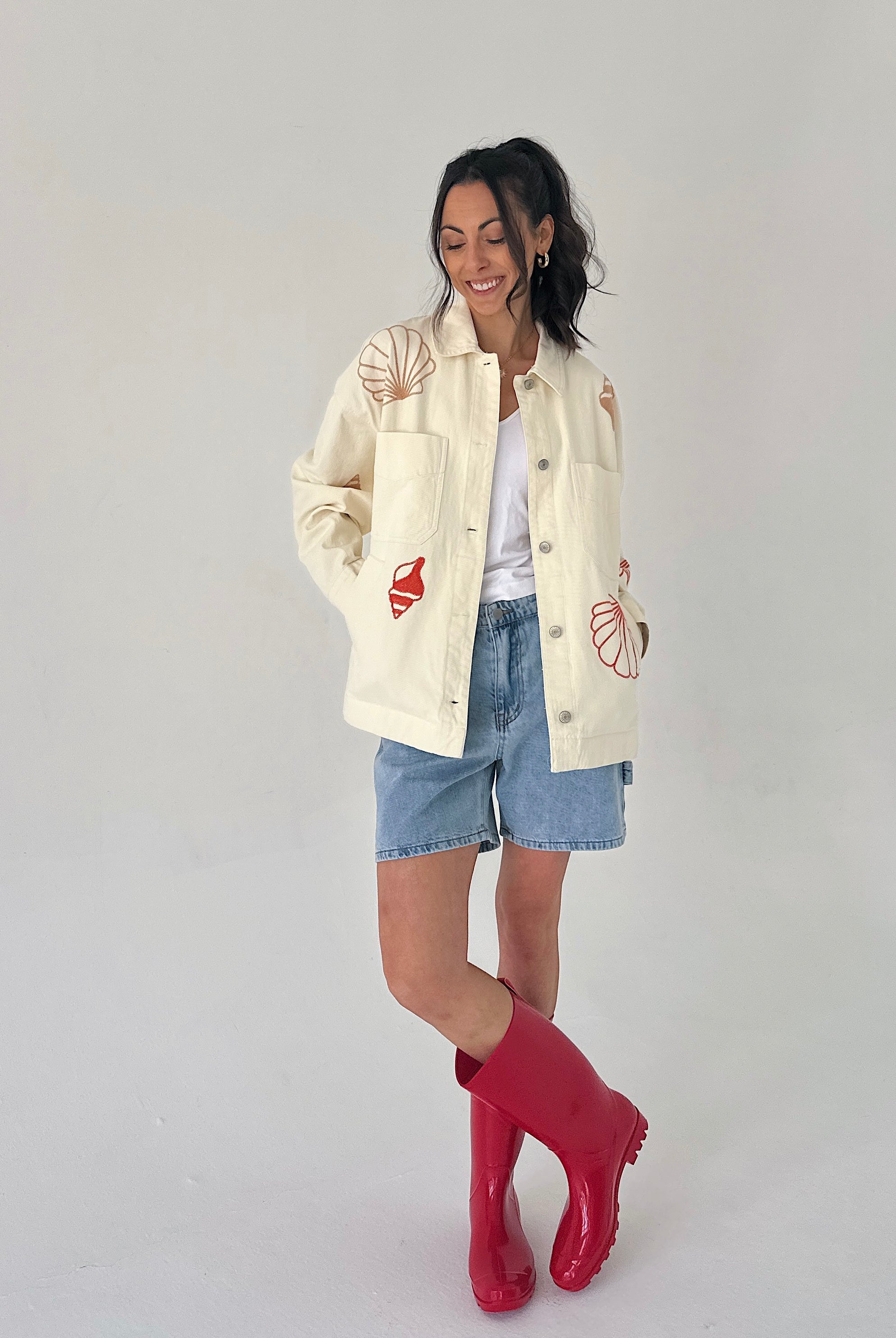 Woman wearing a cream jacket with red and pink seashell designs, denim shorts, and red rain boots on a white background