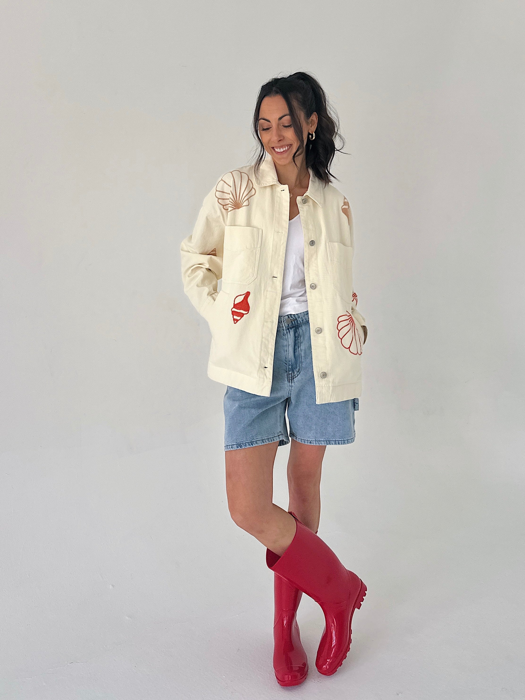 Woman wearing a cream jacket with red and pink seashell designs, denim shorts, and red rain boots on a white background