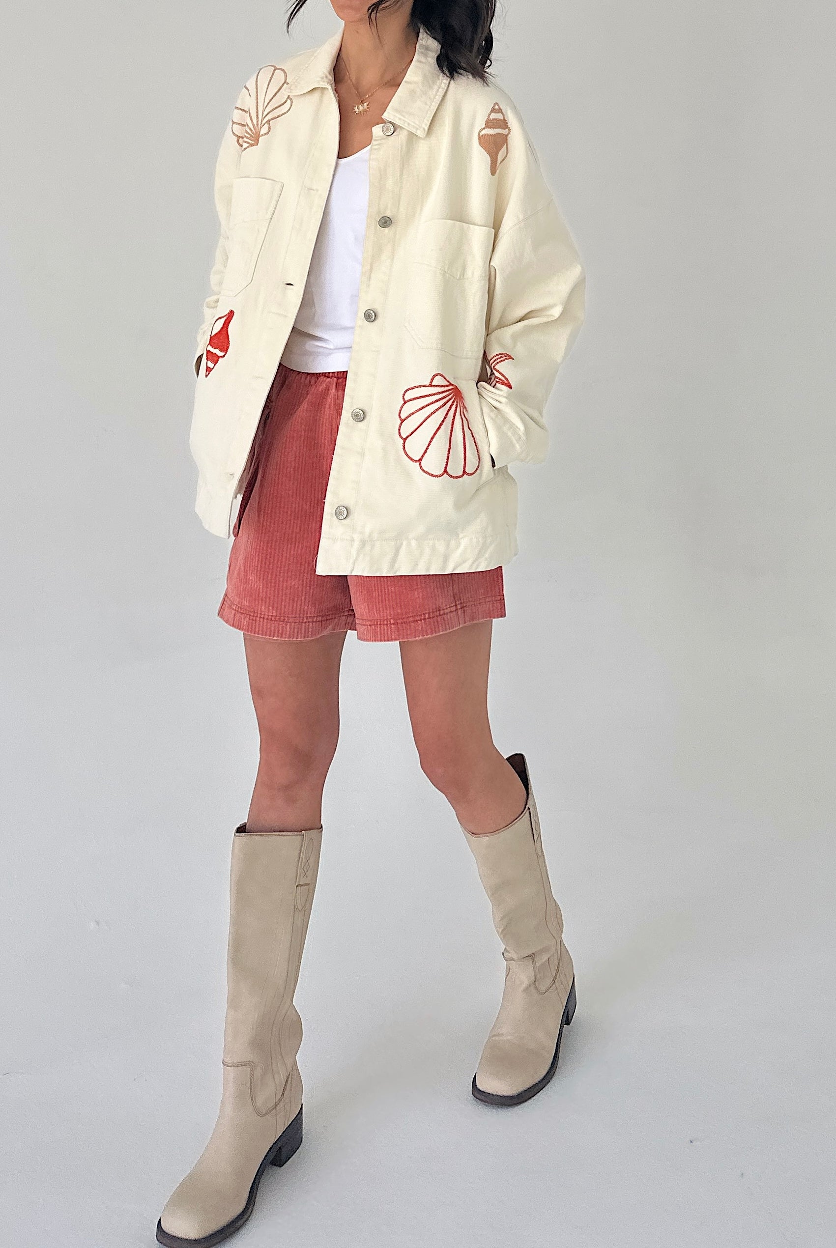 Person wearing a cream jacket with red patterns, rust shorts, and beige knee-high boots on a white background