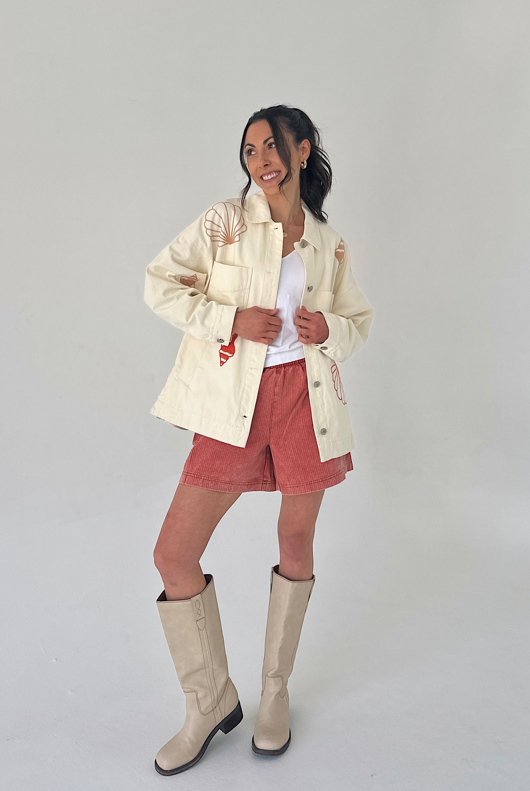 Woman wearing a cream jacket, white shirt, pink shorts, and beige boots on a plain background