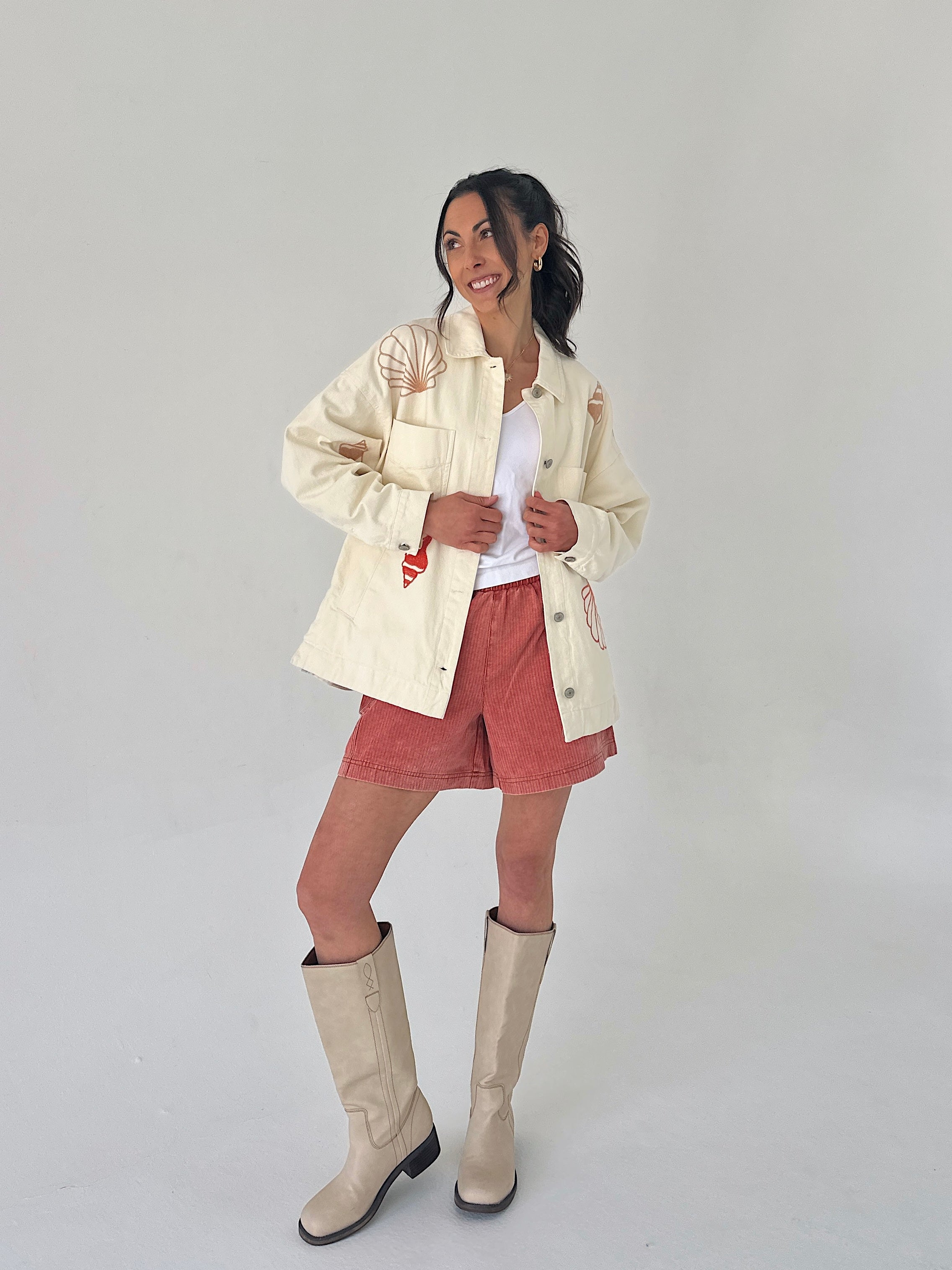 Woman wearing a cream jacket, white shirt, pink shorts, and beige boots on a plain background