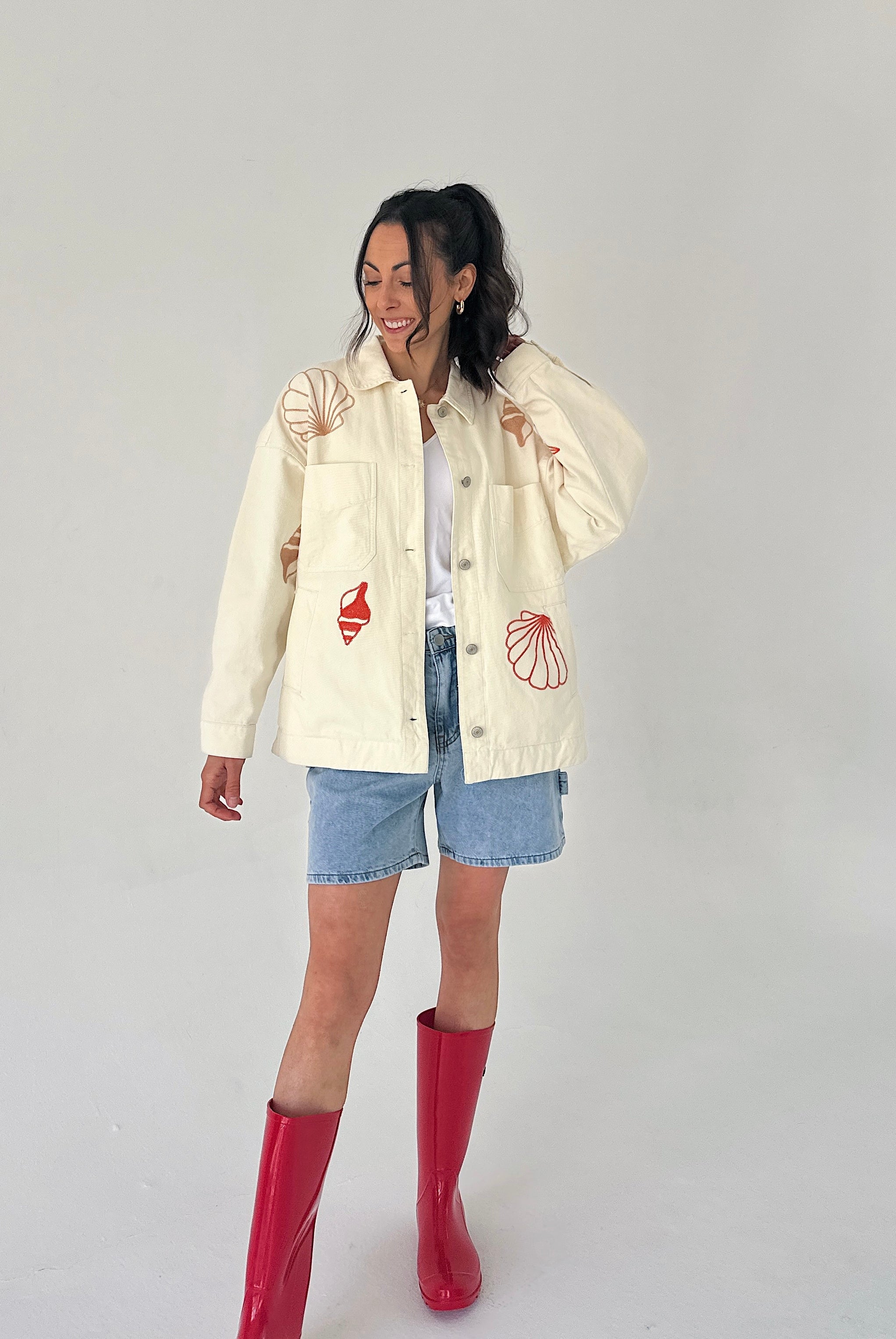 Person wearing a cream jacket with red shell designs, denim shorts, and red boots on a plain background
