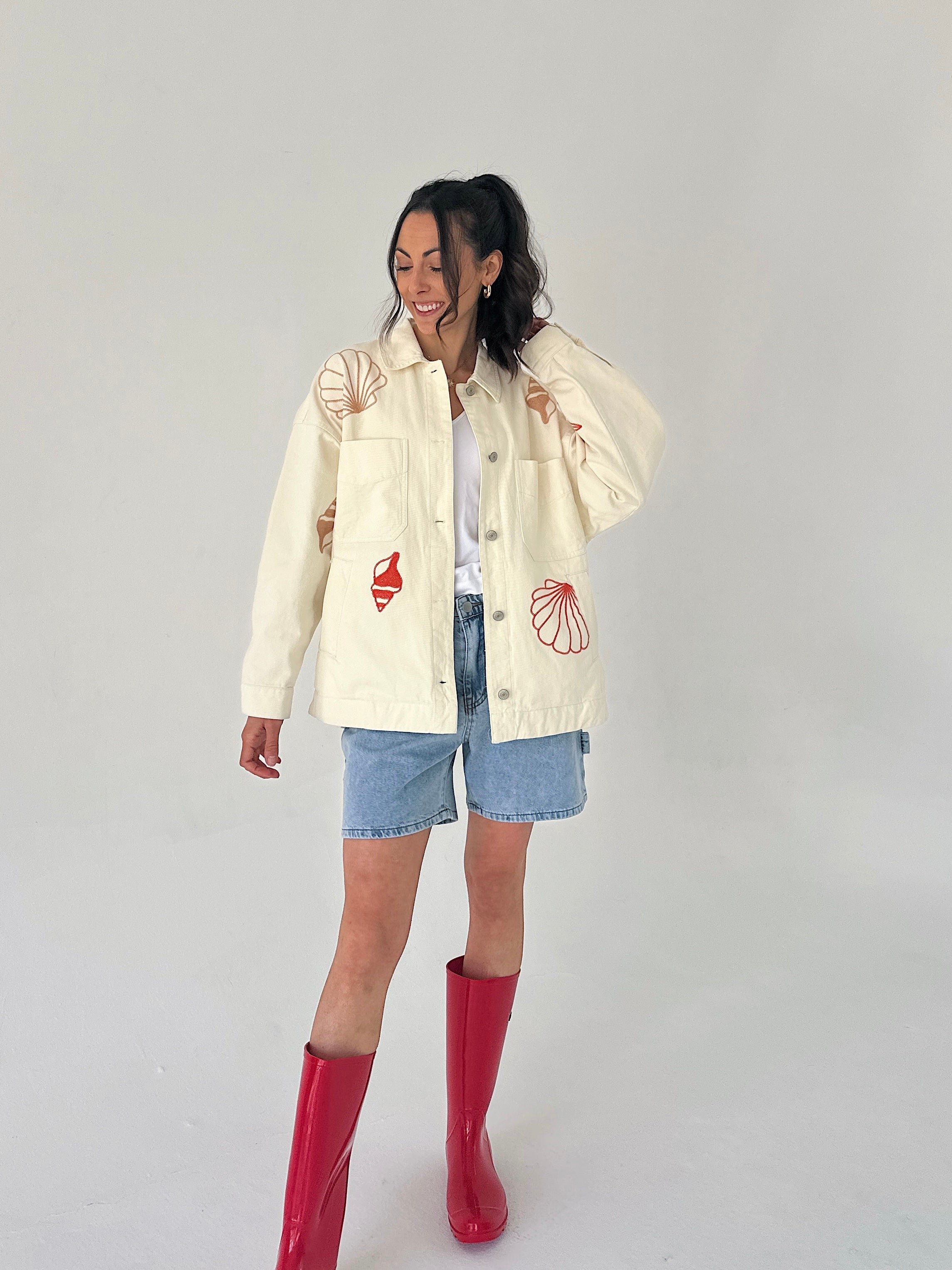 Person wearing a cream jacket with red shell designs, denim shorts, and red boots on a plain background