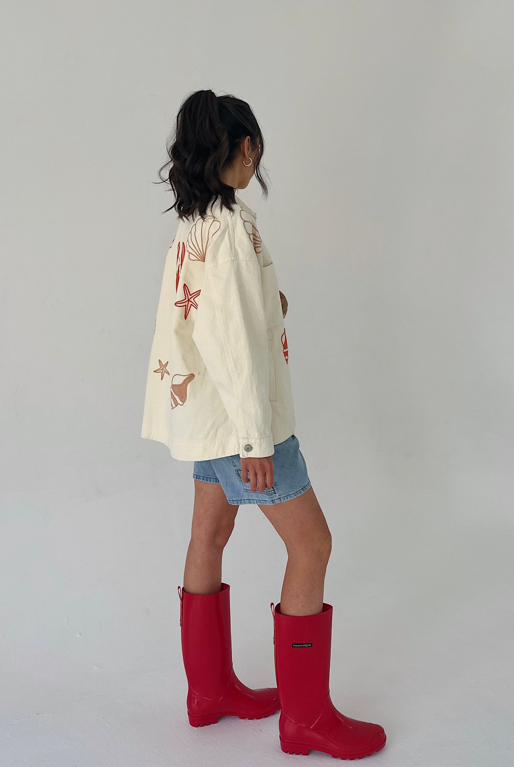Person wearing a cream jacket with starfish and seashell patterns and red rain boots on a plain background