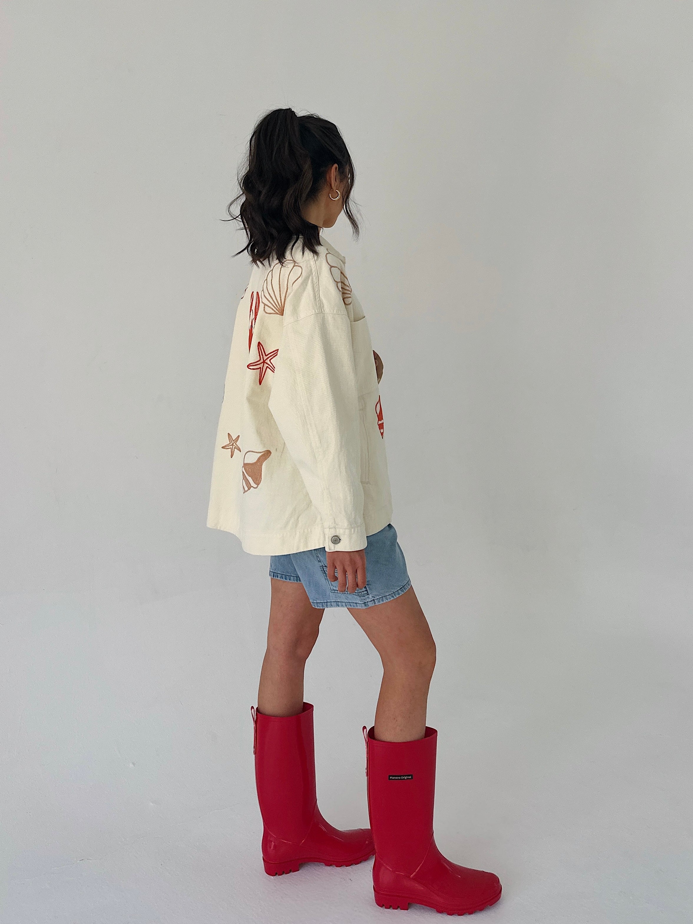 Person wearing a cream jacket with starfish and seashell patterns and red rain boots on a plain background